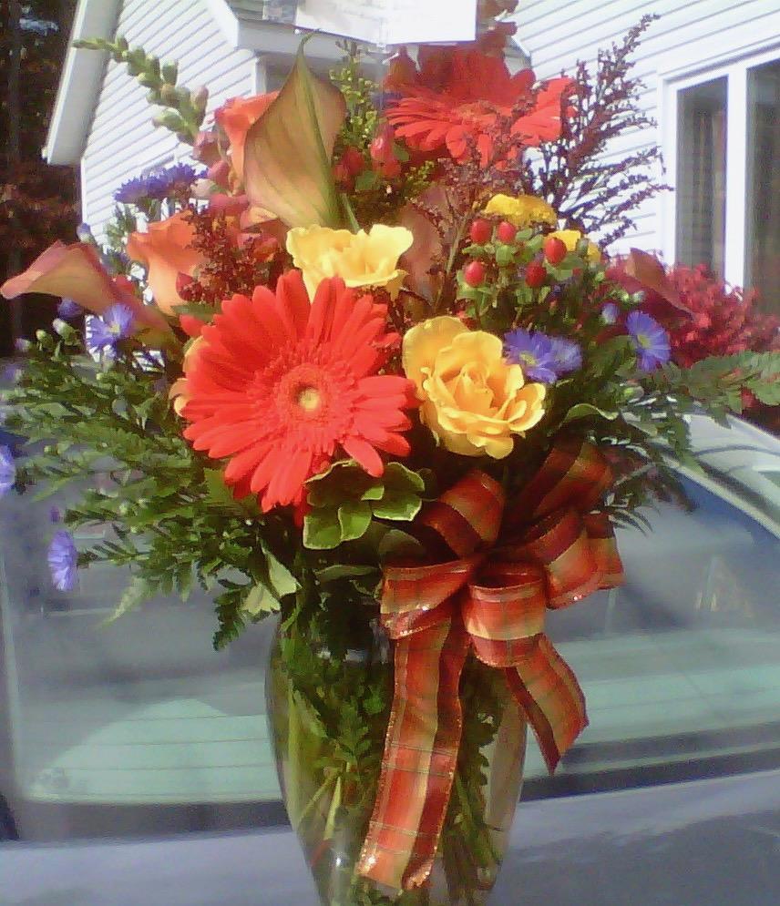 Abundant Fall Flowers Vase - Orange Gerberas, Snap Dragon, Rust colored Calla Lillies, Yellow Roses, Purple Monte, Red Hypericum Berries and  Fall fillers designed in a glass vase with a plaid fall bow