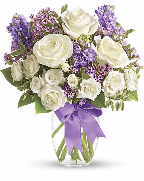 Arrangement of white roses with purple flowers in a clear vase with a lavender bow