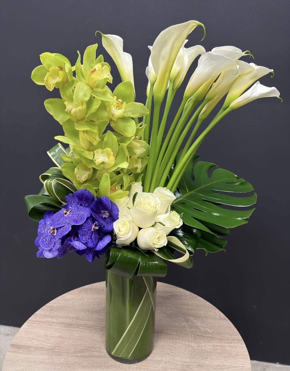 Esmerald Elegance - Green, white, and purple calla lilies, orchids, and roses with glossy monstera leaves in a tall green glass vase-dramatic, polished elegance for upscale events or special gifts.