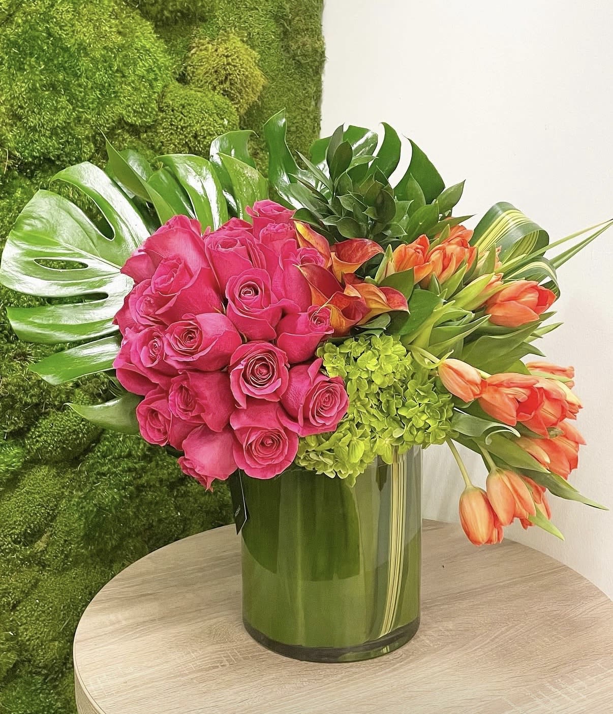 Miami Bloom - Bright pink roses, orange tulips and green hydrangea in a tall green glass vase with lush monstera foliage-vibrant, tropical and celebratory for cheerful gifting or bold displays.