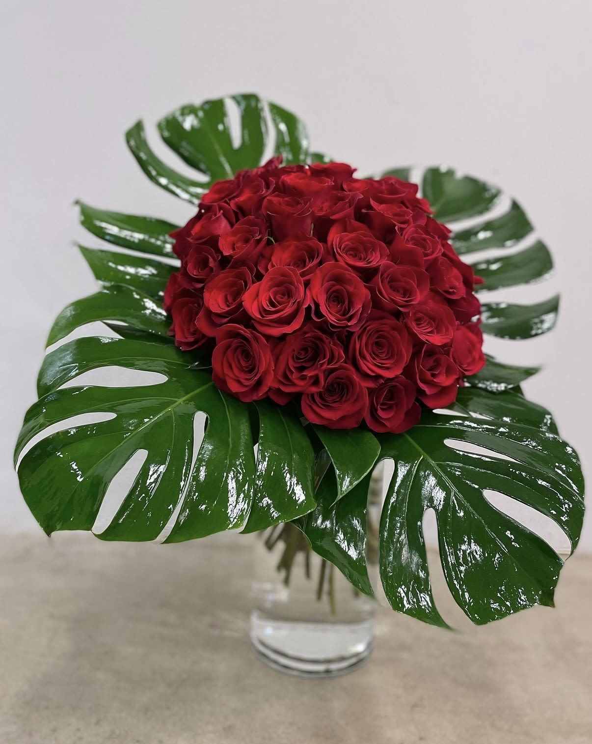 Mon Amour - Red roses with glossy tropical leaves and white accents in a clear glass vase a bold, lush romantic arrangement ideal for anniversaries or love-filled moments.