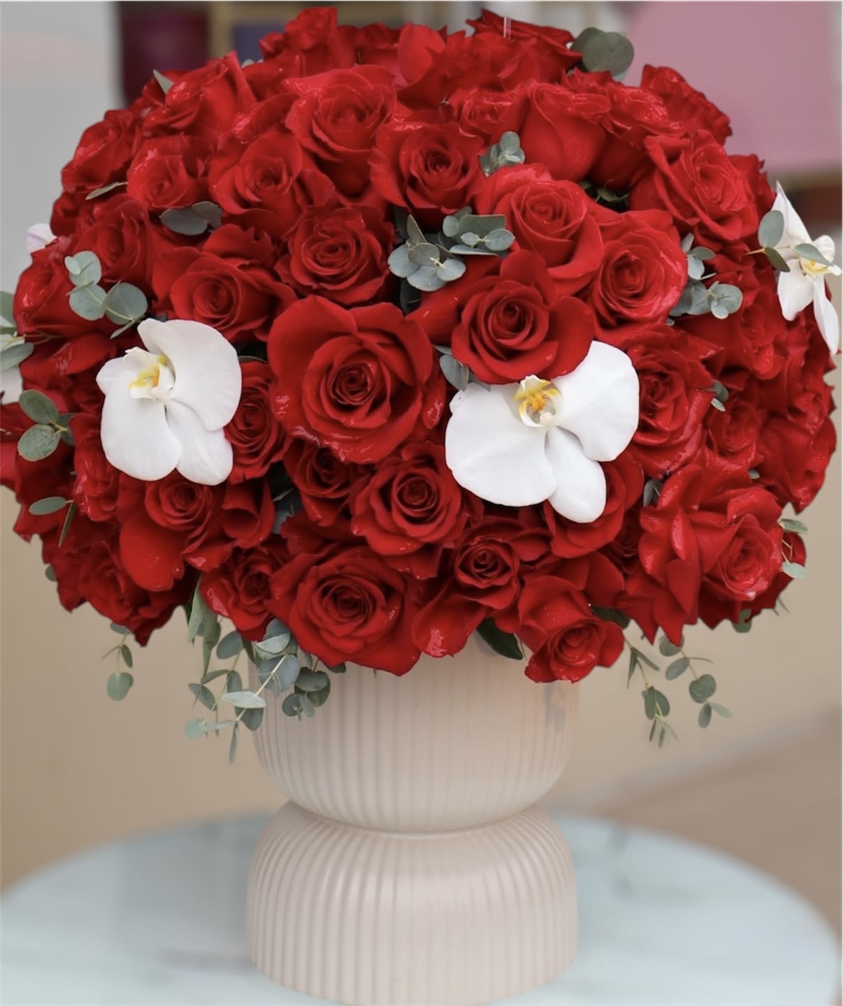 Red Obsession - Rich red roses formed into a velvety dome with white orchid accents and eucalyptus in a ceramic vase - romantic, elegant, and perfect for weddings or love-themed occasions.