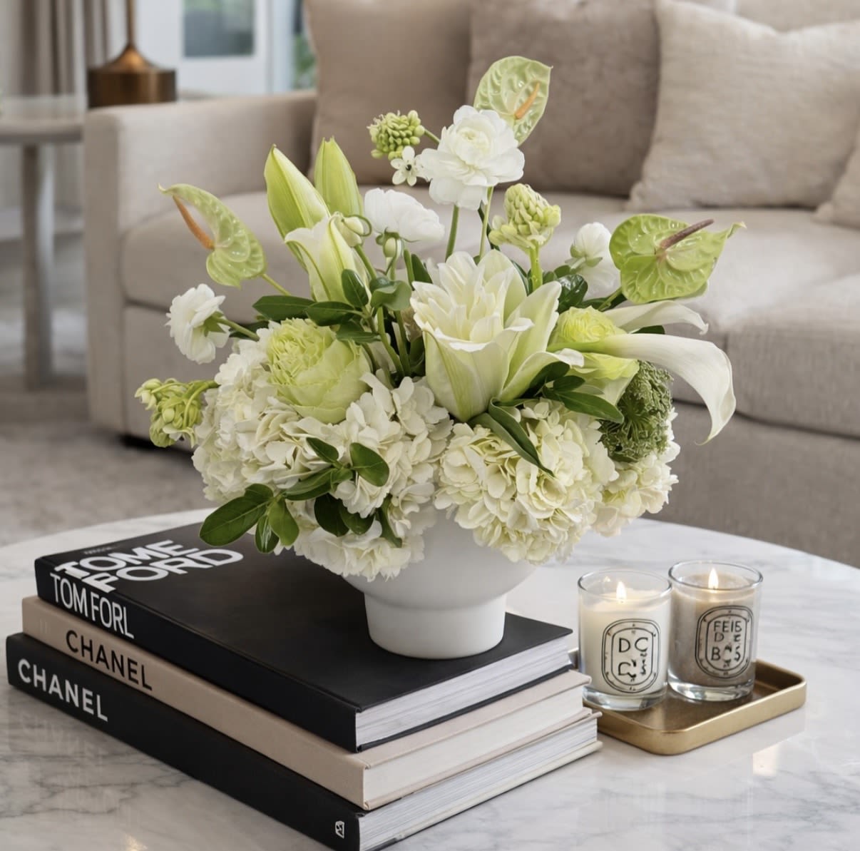 Luna Bloom - White lilies, pale green hydrangea-like blooms and glossy anthurium in a compact white vase-soft, modern palette styled on stacked books with candles for a serene, upscale touch.