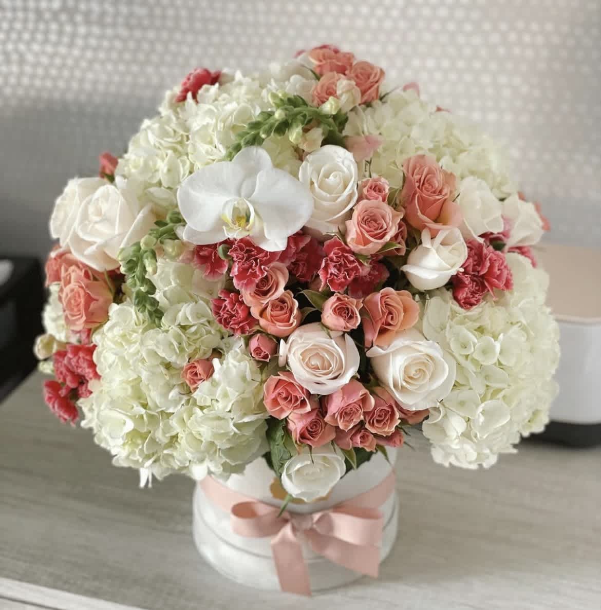 Round bouquet of white hydrangeas, pink roses, and white orchids in a white box with a pink ribbon