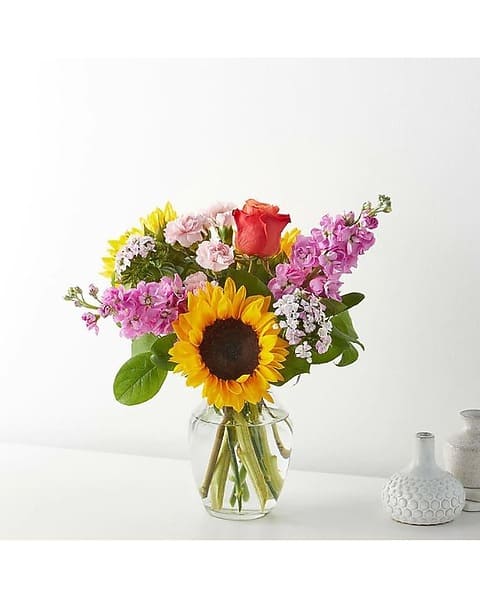 Mixed bouquet with a sunflower, rose, and pink blooms in a clear glass vase