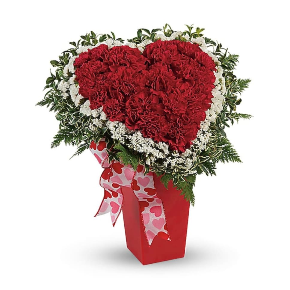 Heart-shaped red carnation arrangement in a red vase with a heart ribbon