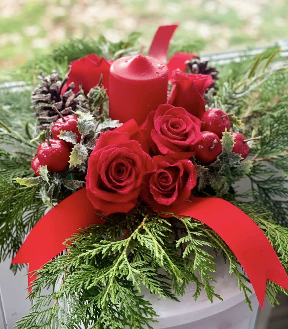 Red roses and a candle arranged with evergreen and berries in a white container.