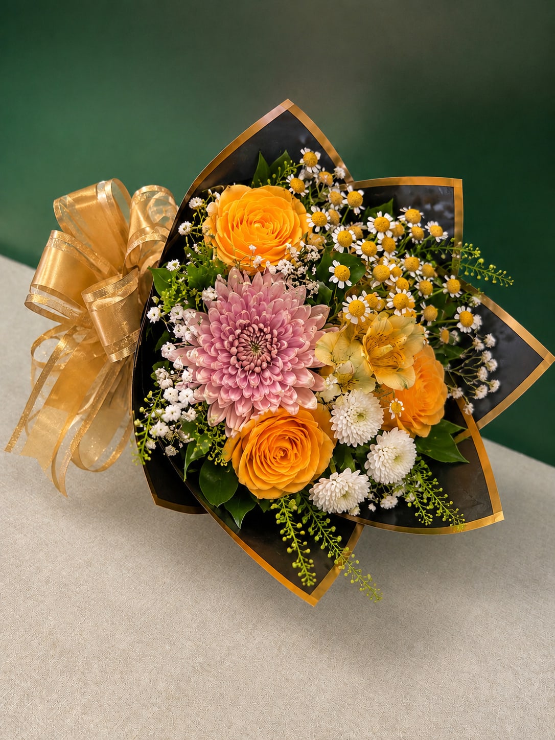 Golden Charm Bouquet Grab &amp; Go - Bright, celebratory and just a little glamorous - the Golden Charm Bouquet Grab &amp; Go is a compact hand-tied bouquet perfect for a birthday or for saying "thinking of you" with warmth. This arrangement blends orange roses and a large pink chrysanthemum up front so the recipient immediately feels seen; it's an excellent choice for Get Well wishes, Graduation cheers, a heartfelt I'm Sorry, or a Just Because surprise that reads as both joyful and refined.  The bouquet mixes alstroemeria and pale yellow alstroemeria, white chrysanthemums, and sunny yellow daisies with the orange roses and the standout pink chrysanthemum for a layered, textured look. Arranged in a rounded, hand-tied style and finished with black wrapping paper edged in gold trim and a large sheer gold ribbon, the contrast of dark wrap and warm florals feels polished and festive. The bouquet's scale is ideal for bedside tables, a dorm room desk, or a small apartment, and the bright pink, white, yellow, and orange palette reads fresh for spring and summer gifting.  Choose this bouquet for moments when you want the person you're thinking of to feel cheered and celebrated - from thank-you gestures and prom bouquets to holiday surprises and romantic notes. Substitutions may be made for seasonal availability while preserving the overall look and value. Fleur De CJ delivers throughout New York, NY, making it easy to send prompt, thoughtful flowers that make someone's day.