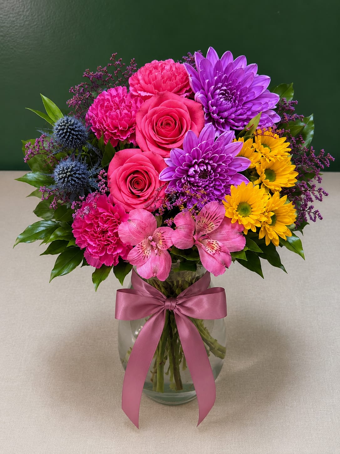 Colorful bouquet of roses, chrysanthemums, carnations, and alstroemeria in a glass vase with a pink ribbon