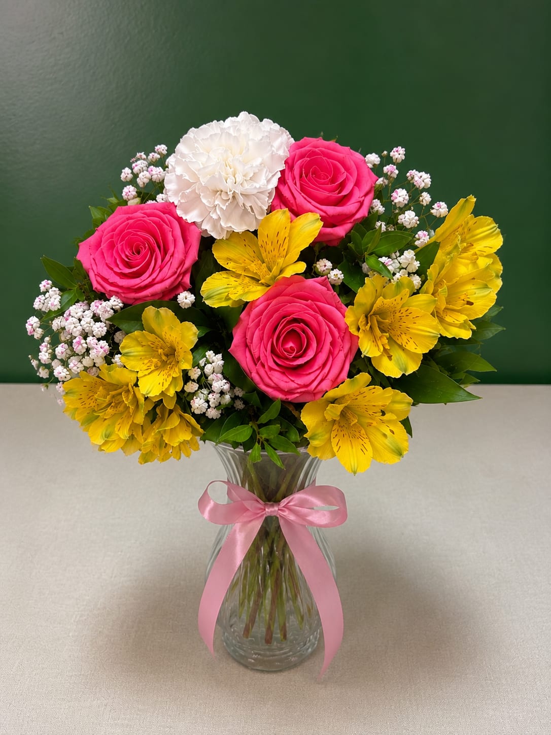 Pink roses, white carnation, and yellow alstroemeria in a glass vase with a pink ribbon