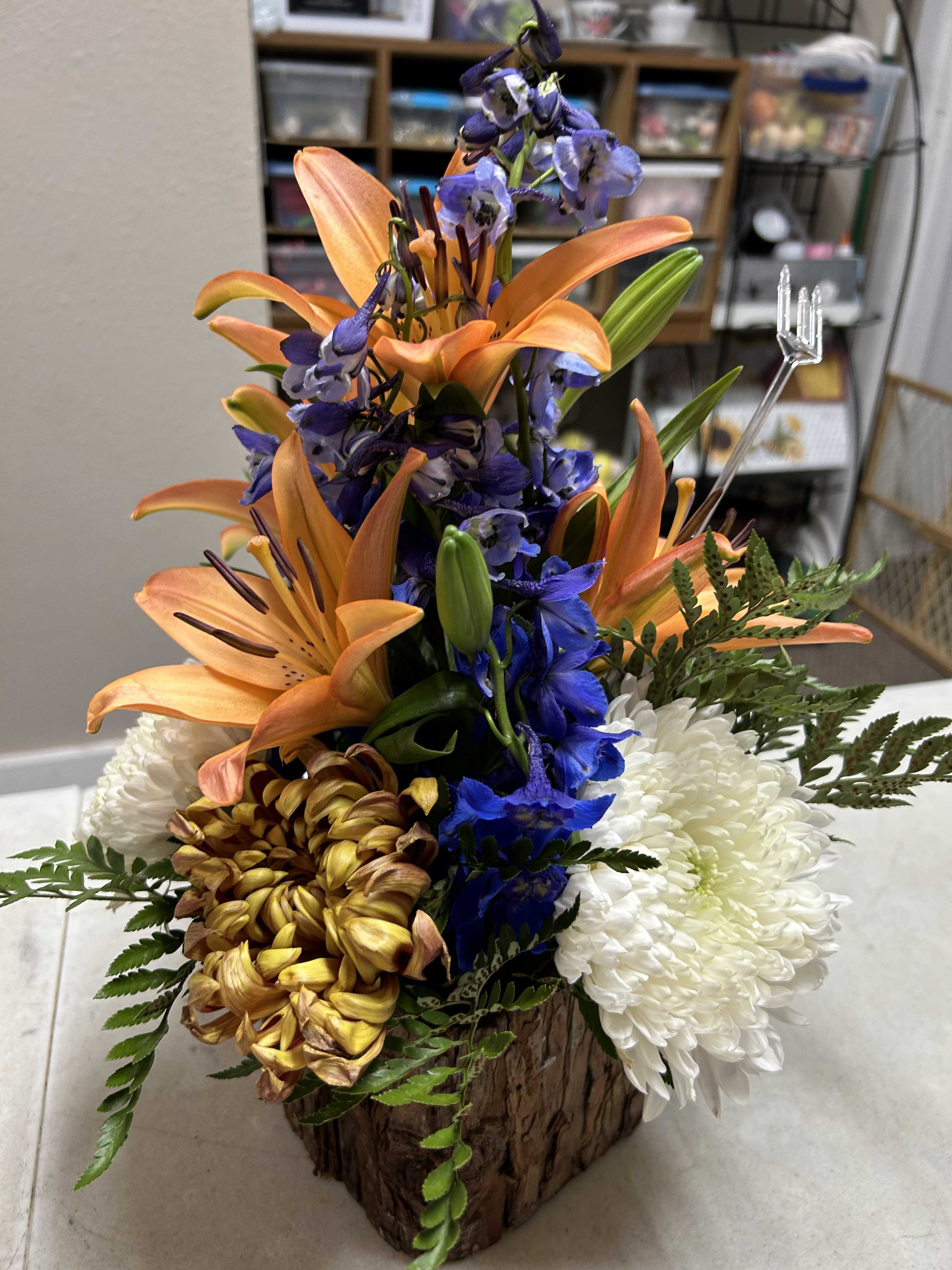 Orange lilies with blue delphinium and white chrysanthemums in a wood-look container