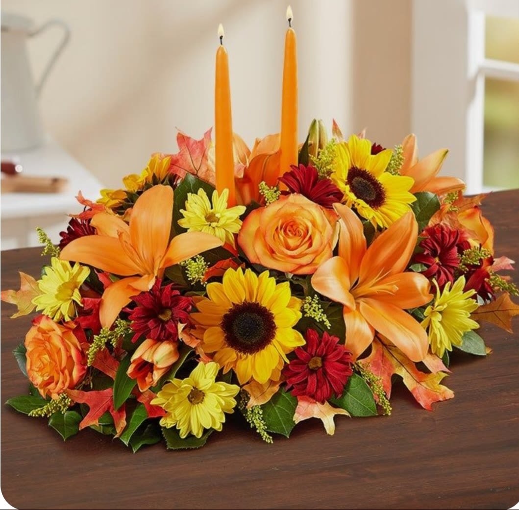 Vibrant fall centerpiece  - A vibrant fall centerpiece featuring two tall, orange candles surrounded by an arrangement of autumn flowers. The floral display includes bright orange lilies and roses, sunny yellow poms, and red hypericum berries, all set against a backdrop of lush green leaves and scattered autumn foliage. This stunning arrangement is perfect for adding a touch of seasonal warmth to any table setting