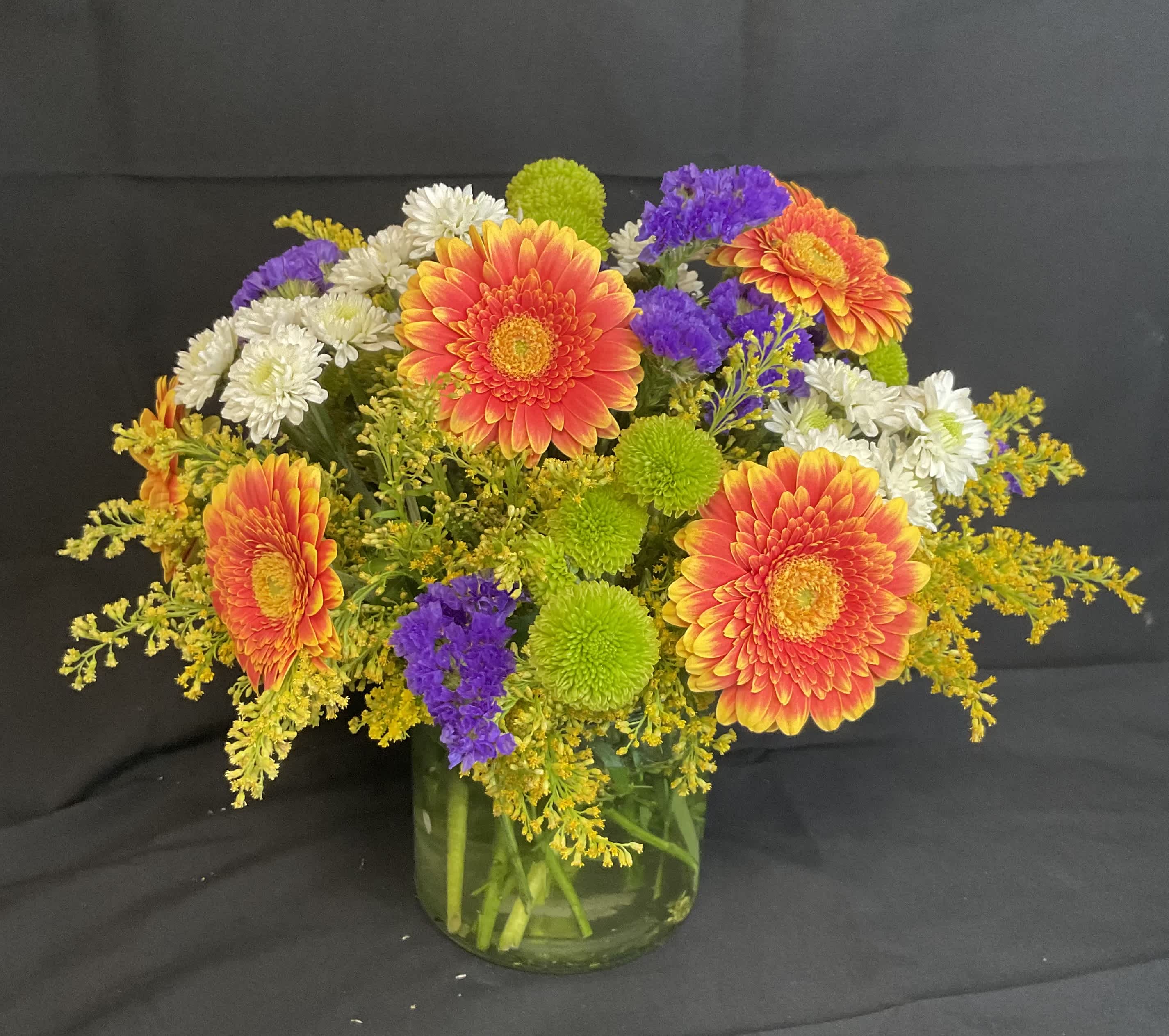 Mixed bouquet of orange gerbera daisies, white mums, and purple filler in a glass vase