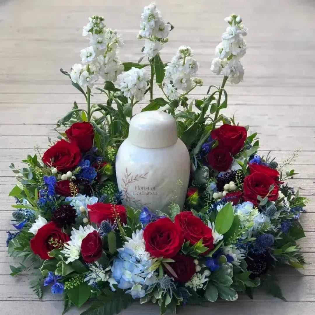 Red roses and white flowers arranged around a white urn on a wooden surface.
