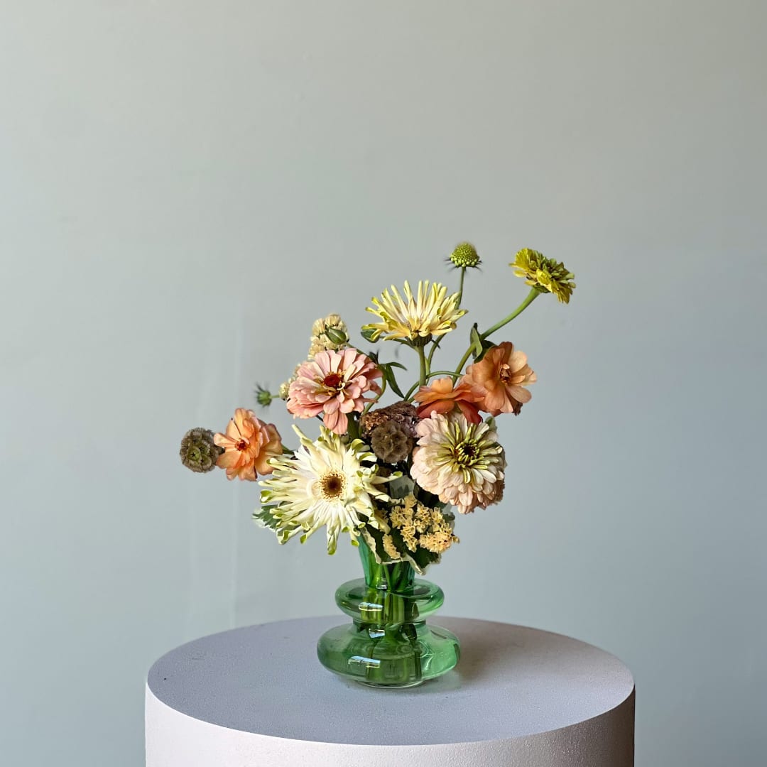 Mixed pastel flowers in a green glass vase on a round pedestal