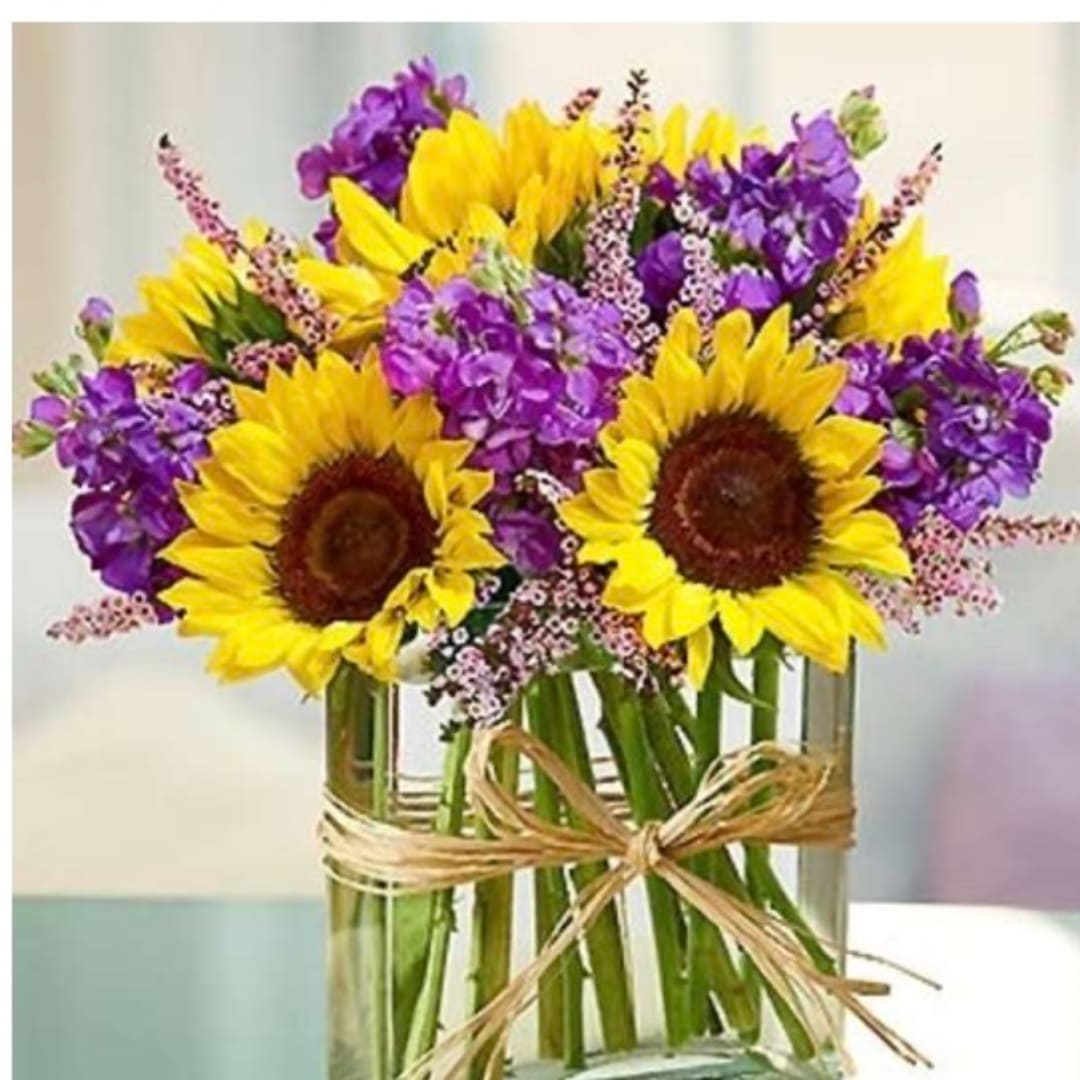 Sunflowers and purple flowers in a clear glass vase with a raffia bow