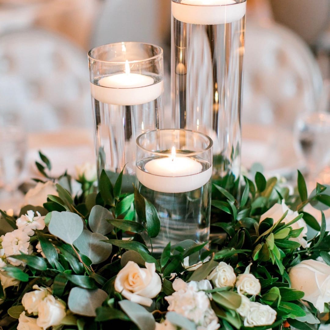 White roses and greenery surround three glass candle cylinders