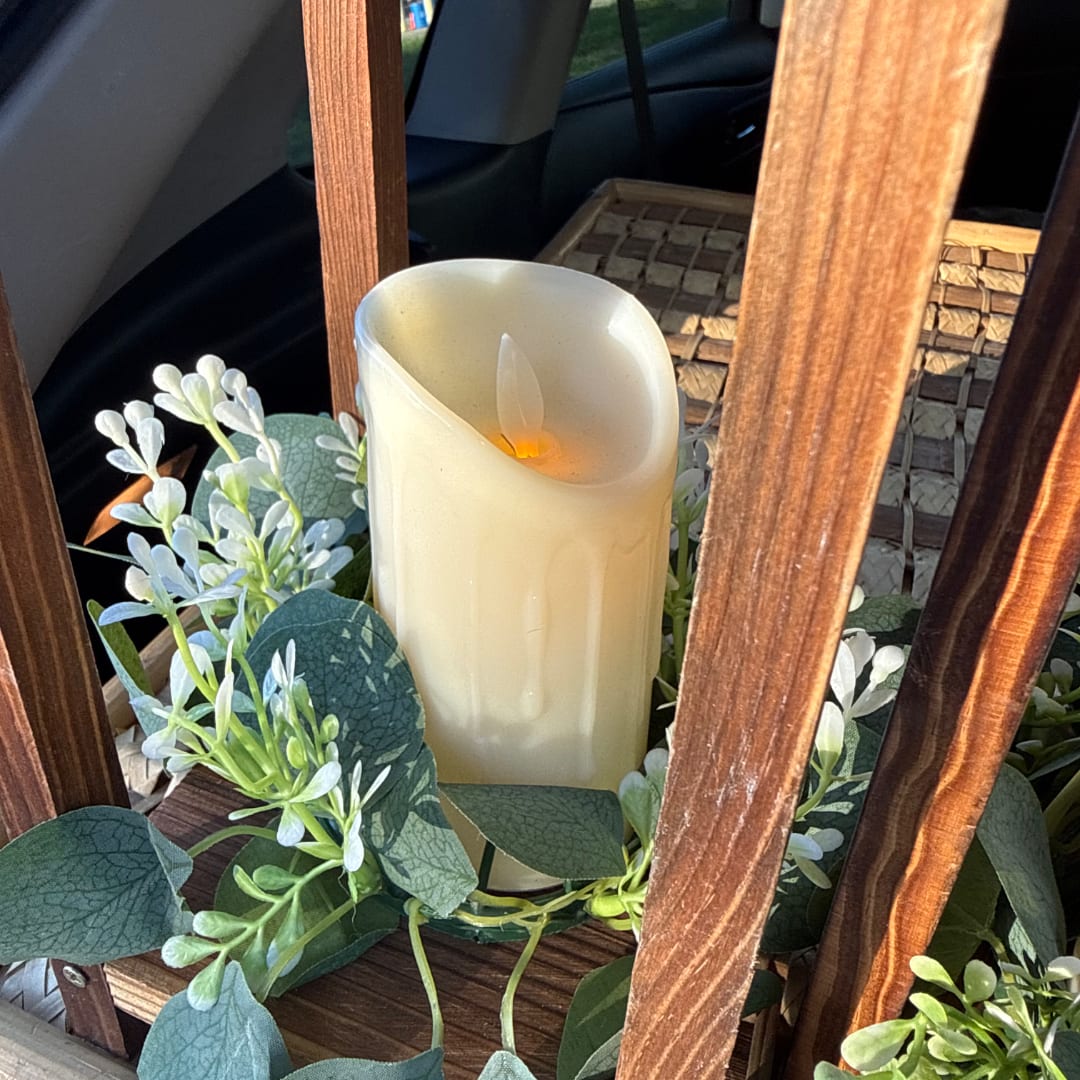 Cream pillar candle surrounded by white flowers and green leaves in a wooden lantern