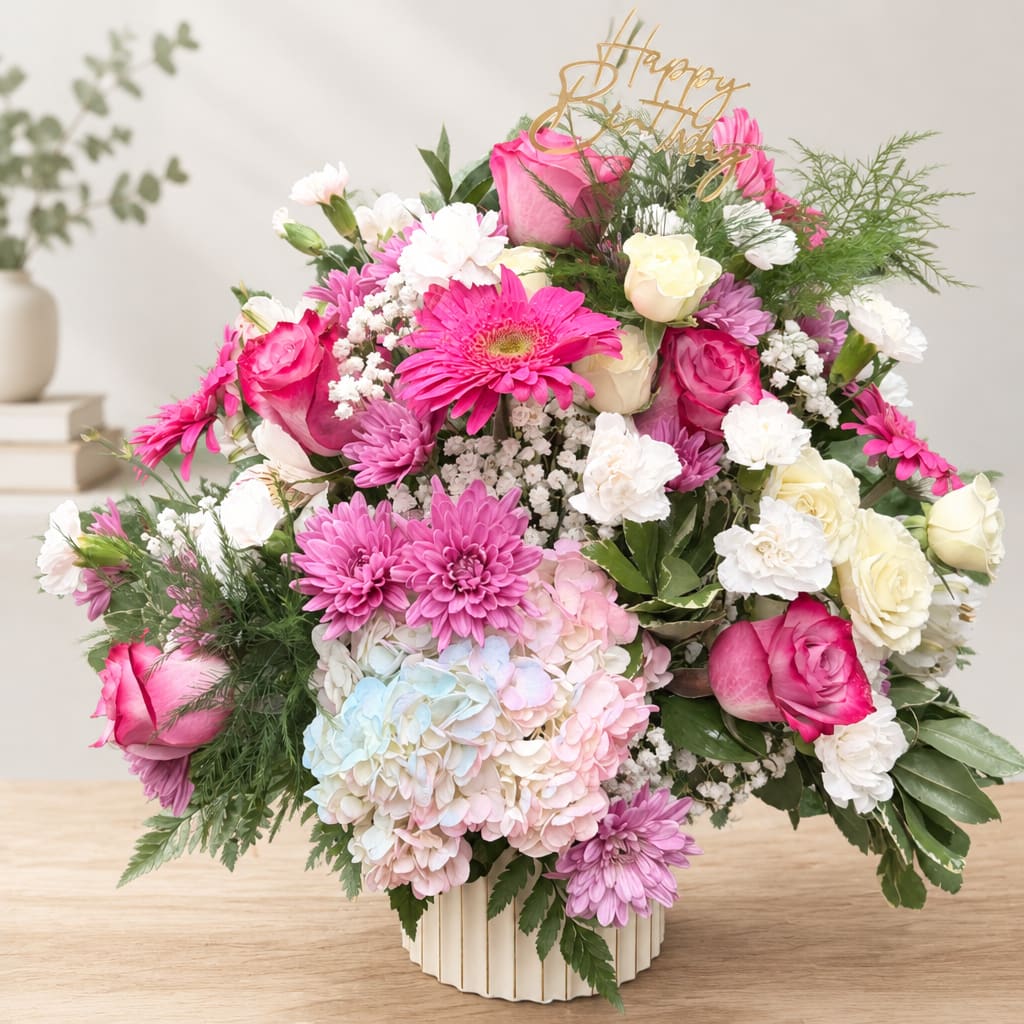 Pink Rose and Hydrangea Birthday Bouquet - A lush mix of bright pink roses and pastel hydrangeas that reads like a party in bloom - the Pink Rose and Hydrangea birthday bouquet is designed to make her feel truly celebrated. This birthday flower arrangement pairs vivid pink roses and classic white roses with pink and lavender daisy-like blooms and white carnations, so the recipient sees both bold color and gentle pastels at once; a gold "Happy Birthday" topper adds a festive finishing touch she'll notice the moment the box is opened.  Carefully rounded and full, the design sits in a ceramic vase and uses airy filler and feathery greenery for soft, layered texture that feels both feminine and joyful. The centerpiece hydrangea cluster gives volume while the roses and daisies provide pops of pink, purple, and white for a mixed-color effect that's ideal for moms, sisters, friends, or coworkers. Arrives ready to display in Dublin, OH - choose your size to confirm vase dimensions, and refresh with a daily water change to keep blooms lively throughout the celebration.