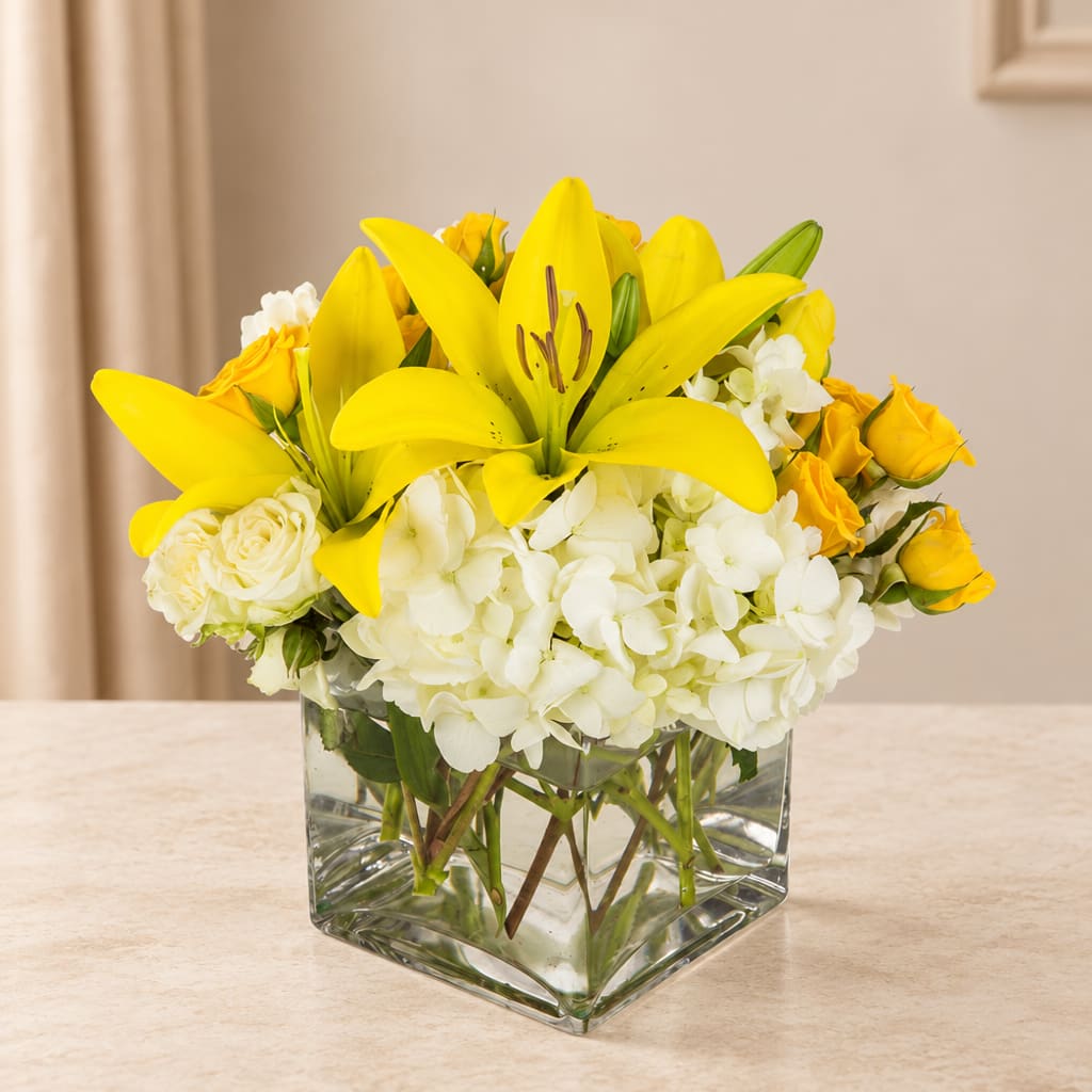Yellow lilies and white flowers arranged in a clear square vase