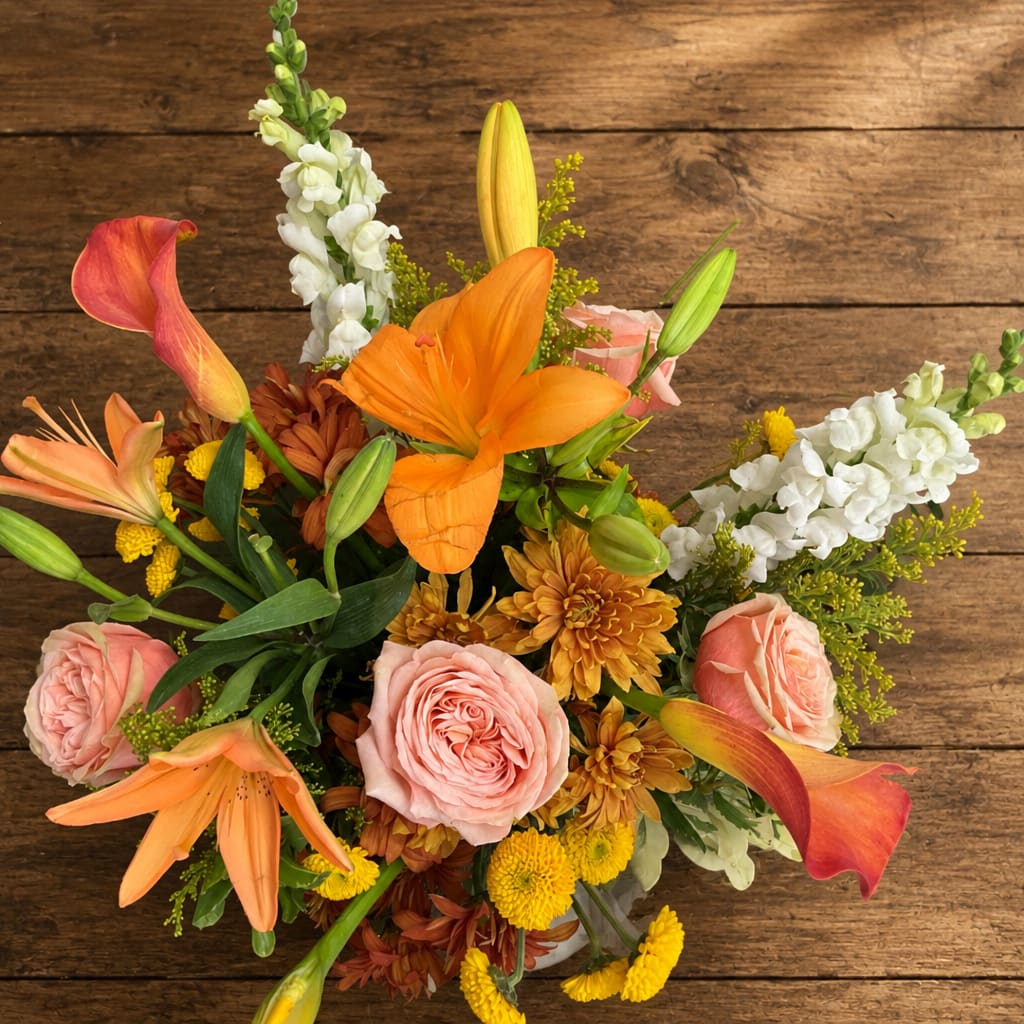 Orange Lily and Pink Rose Arrangement in White Bucket - Bright orange lilies take center stage in this joyful flower arrangement, making it an ideal birthday bouquet or congratulations gift that will make the person you're thinking of feel truly celebrated. The Orange Lily and Pink Rose Arrangement in White Bucket pairs the vivid energy of orange lilies with soft pink roses to create a warm, cheerful statement-perfect for springtime surprises, a festive table accent, or an autumnal pick-me-up for someone special in Romeoville, IL.  This hand-arranged mix is full and textural: bright orange lilies and pink roses are complemented by white snapdragons, yellow and bronze chrysanthemums, and occasional calla lilies for contrast. Stems are gathered tightly and presented in a simple white bucket (also shown in photos with alternate clear glass vases); the composition reads lively and slightly asymmetrical, with lily buds and blooms extending outward for an airy, celebratory silhouette. Colors include orange, pink, white, yellow and green for a vibrant, layered look.  Send this arrangement to mark a birthday, say thank-you, or celebrate a milestone-the recipient will feel noticed and cheered by its bold, sunny palette. Sizes and vessel options shown in photos display slight variations (white bucket or clear/green glass vase); for best longevity, trim stems and change the water every couple of days to keep blooms fresh.