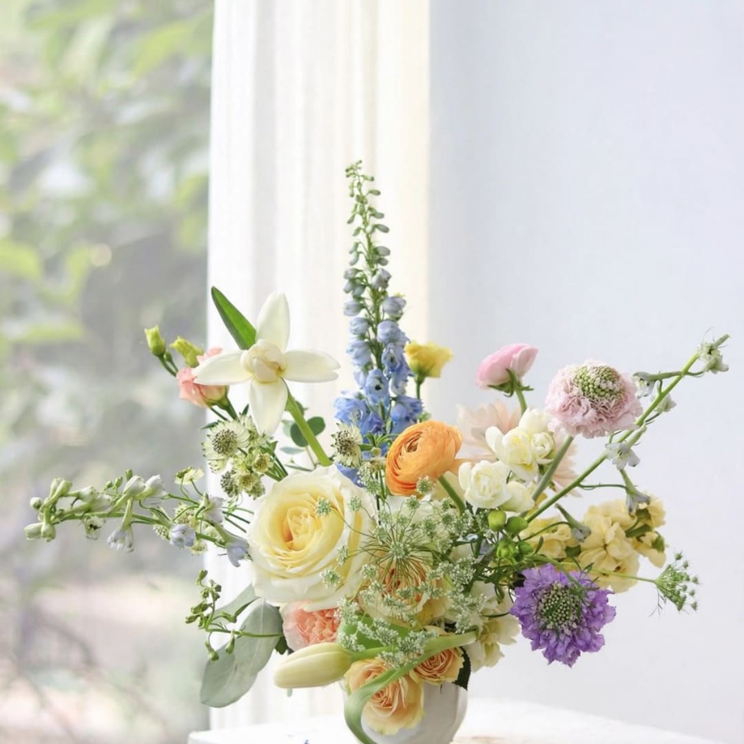 Mixed pastel flower arrangement in a white vase by a window