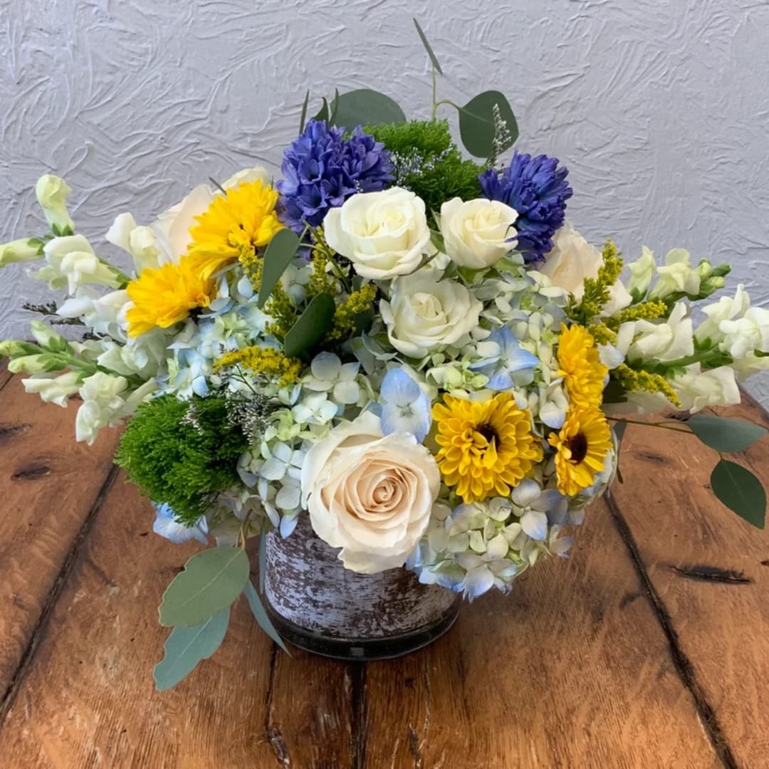 Mixed bouquet in a rustic metal container with white roses, yellow blooms, and blue flowers