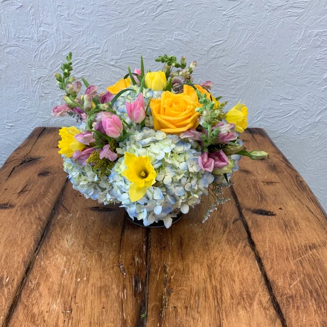 Mixed bouquet with yellow roses, pink tulips, and blue hydrangeas in a low container
