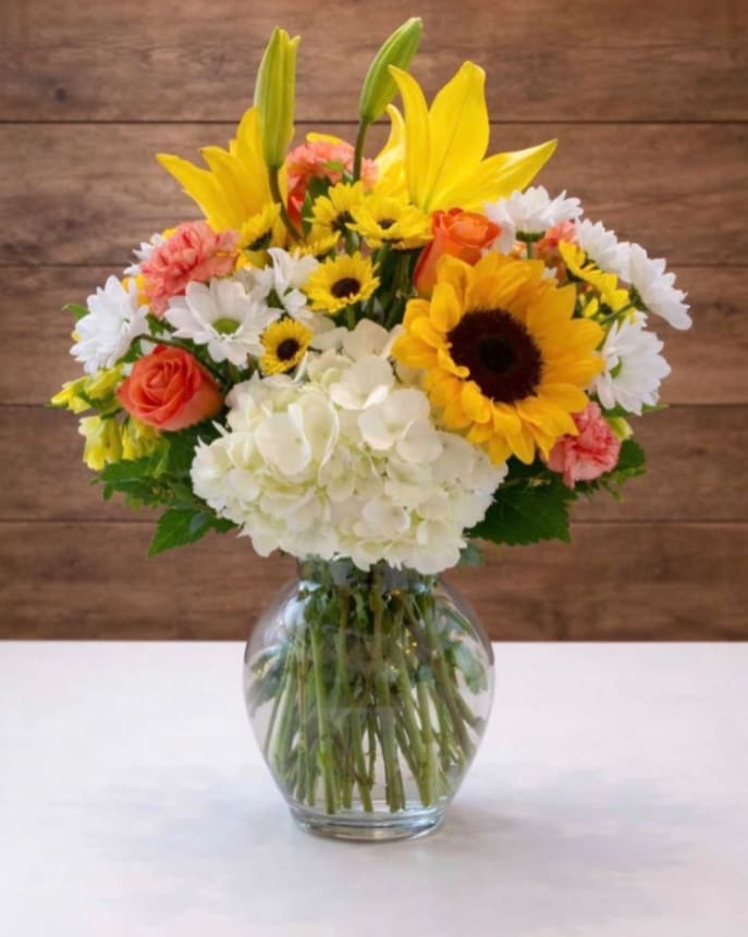 Mixed bouquet of yellow lilies, sunflowers, roses, daisies, and hydrangea in a glass vase