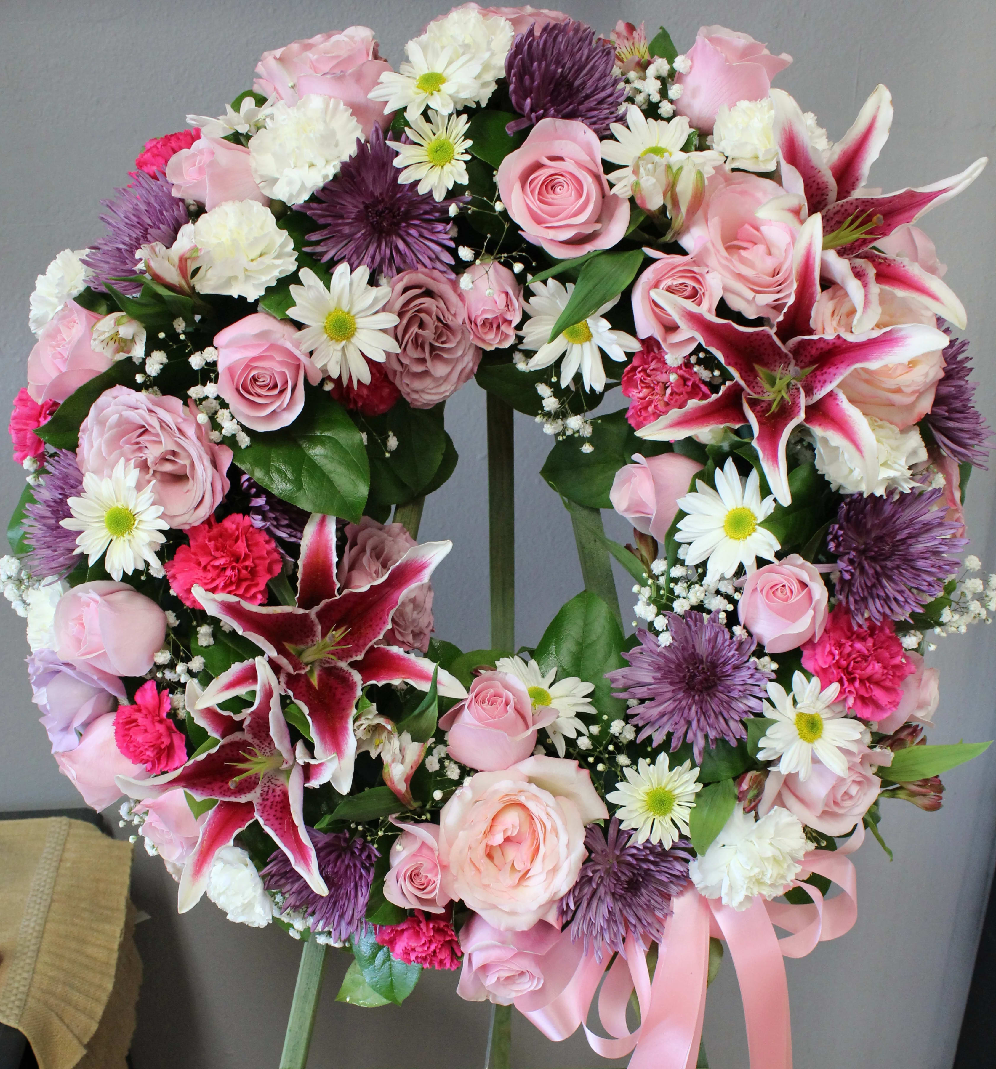 Standing wreath of pink roses, lilies, white daisies, carnations, and purple mums with a pink ribbon on an easel