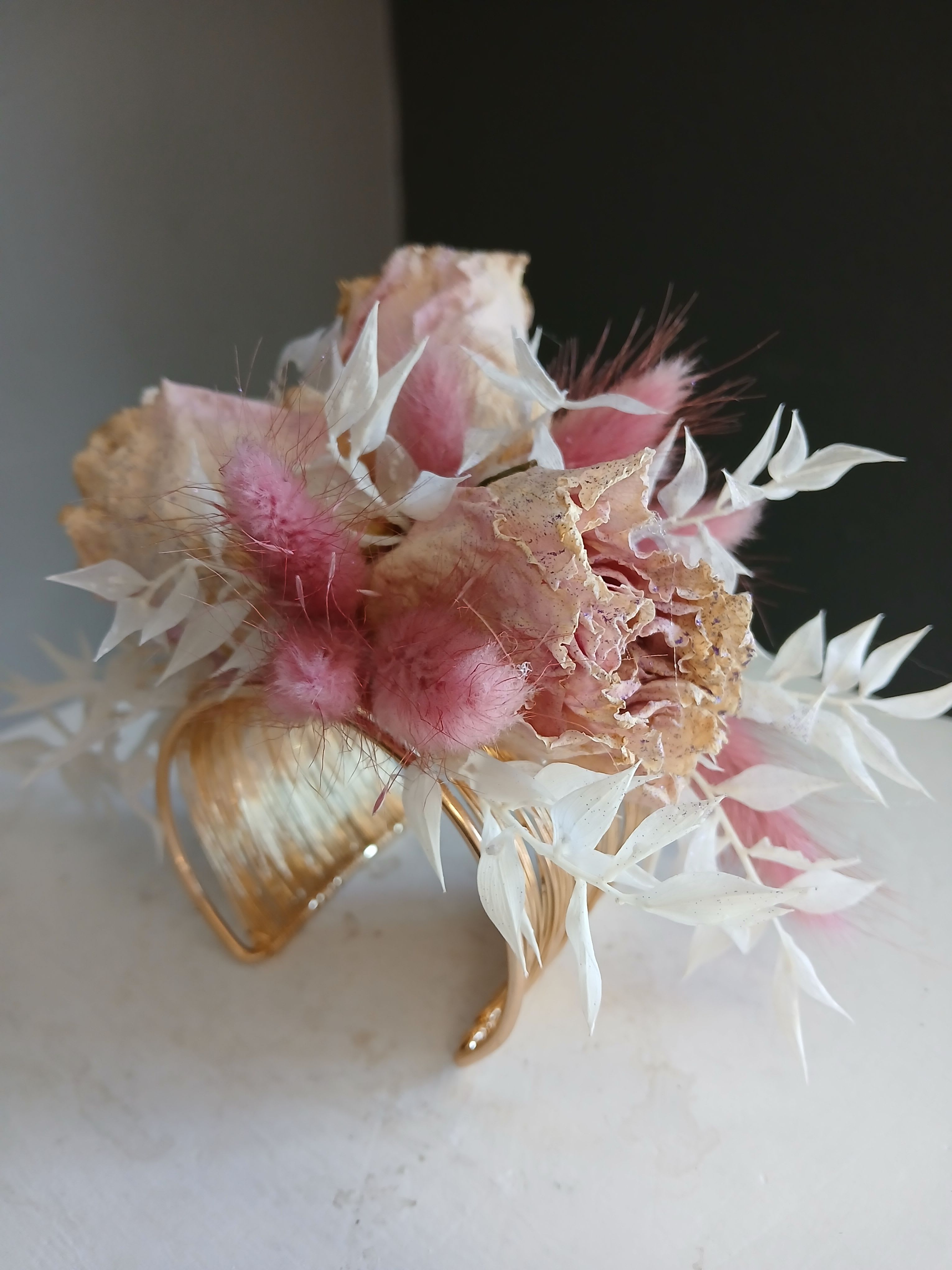 Dried corsage Pinky Blush gold band - A petite dried corsage in soft pink and white that feels like a keepsake.  The cluster is made of papery, textured dried blooms and airy white accents, tightly grouped for a compact, handcrafted silhouette. The gold metal holder frames the composition and gives it an elegant, vintage-inspired finish that catches the light and elevates the soft, blushed palette.  Ideal for the person who favors lasting, low-maintenance florals rather than fresh stem. Because it's dried, no water is needed; keep it out of direct sunlight and it will retain its color and shape as a long-lasting memento.