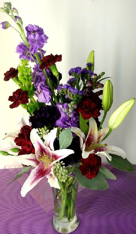 Mixed bouquet with lilies, purple blooms, and red carnations in a glass vase