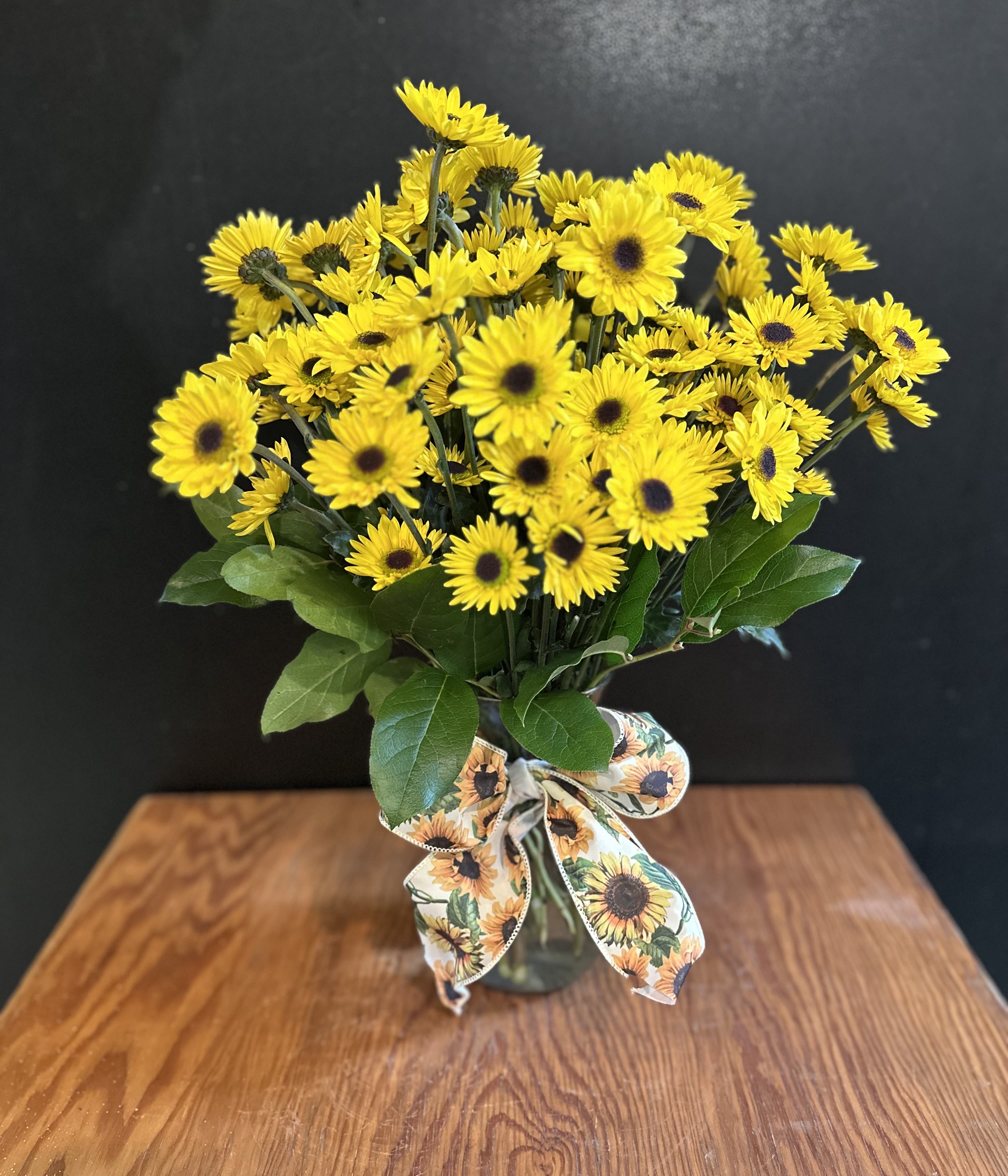 Yellow daisy bouquet in a glass vase with a sunflower-patterned ribbon