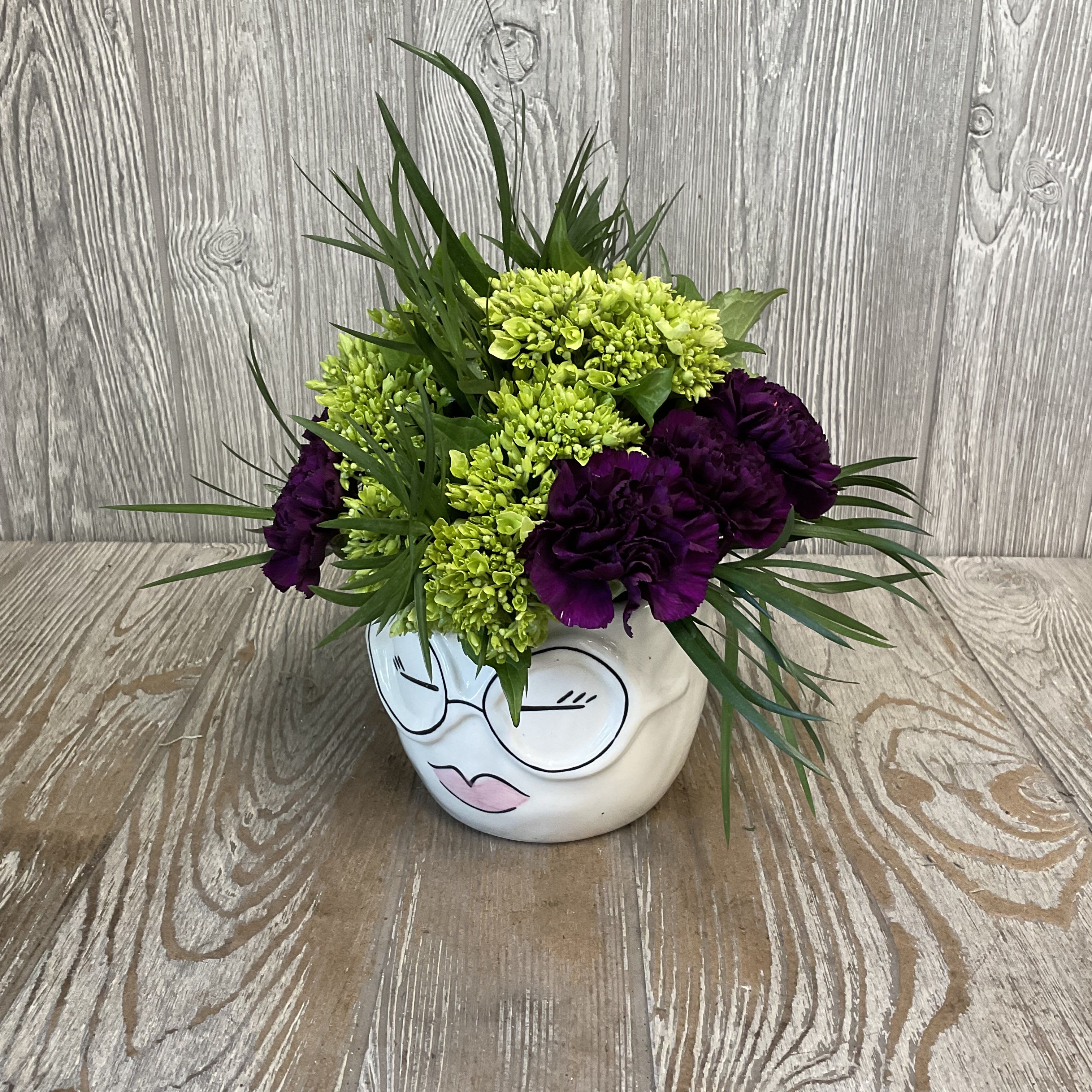 Purple and green floral arrangement in a white face-shaped vase
