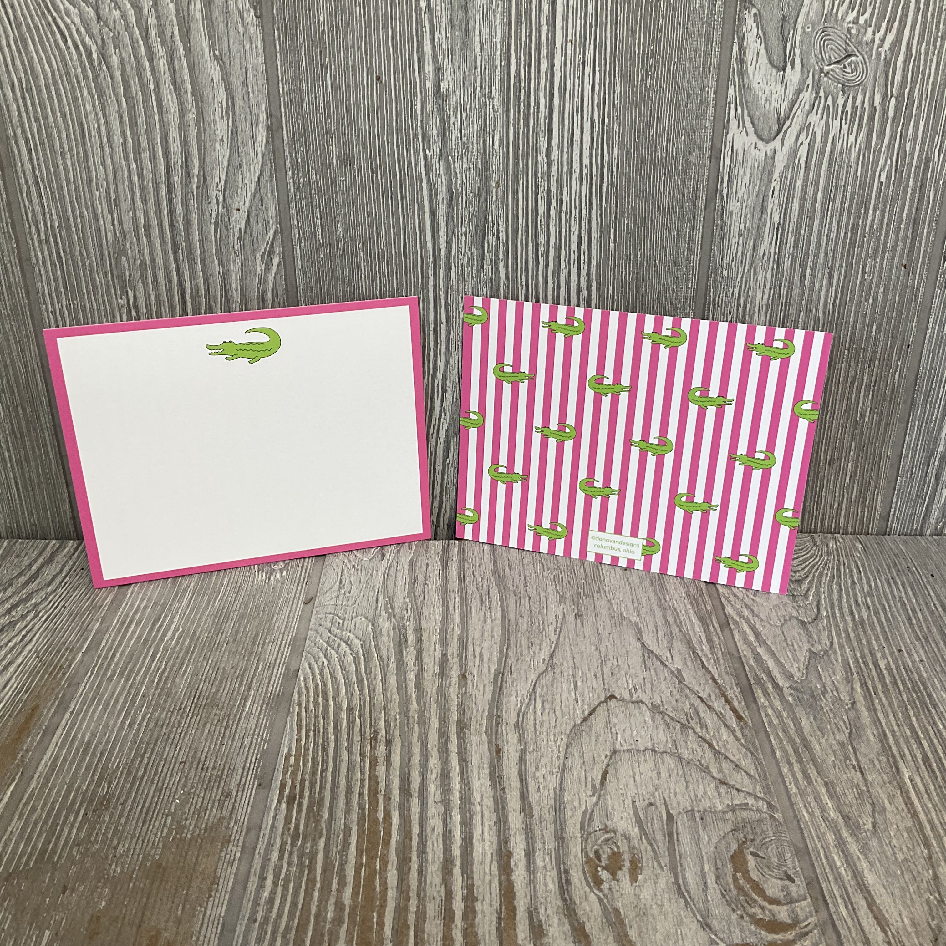 Gator Flat Notecards with Envelopes by Donovan Designs - Bright, playful notecards perfect for everyday messages and casual gifting - these flat stationery cards pair a bold pink border with a cheerful green alligator motif for a fresh, upbeat look. Use them as birthday notes, party thank-yous, or simple just-because messages when you want the recipient to smile; the design reads youthful and friendly rather than formal, so they're ideal for kids, friends, or anyone who appreciates a whimsical touch.  Each set includes 10 flat greeting cards with coordinating white envelopes, packaged in clear plastic for a retail-ready presentation. The card front shows a clean white face framed in bright pink with a small green alligator near the top; the back features pink-and-white vertical stripes with repeated alligator prints. Lightweight and easy to mail or tuck into a bouquet, these notecards add a playful accent to gifts and arrangements.