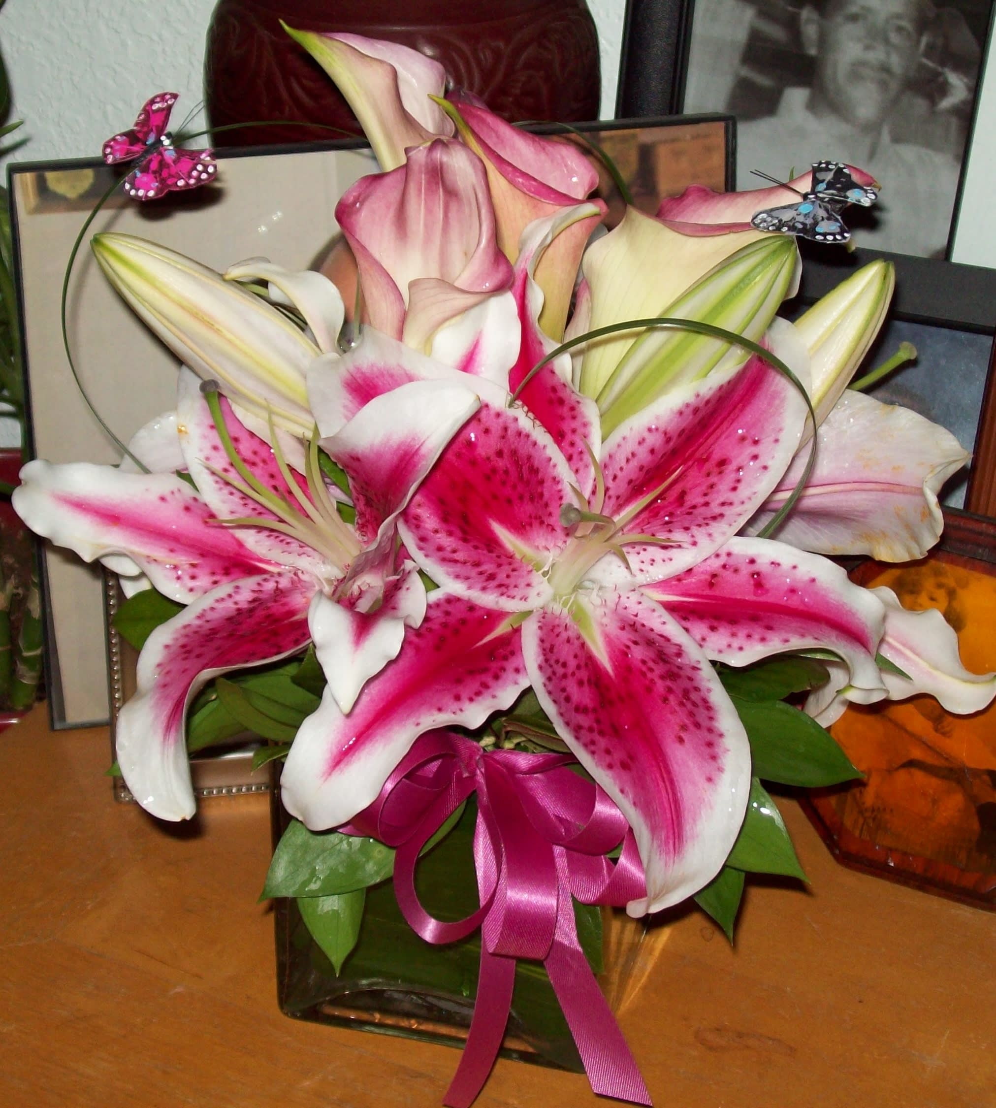 Pink and white lilies arranged in a glass vase with a ribbon and butterfly picks.