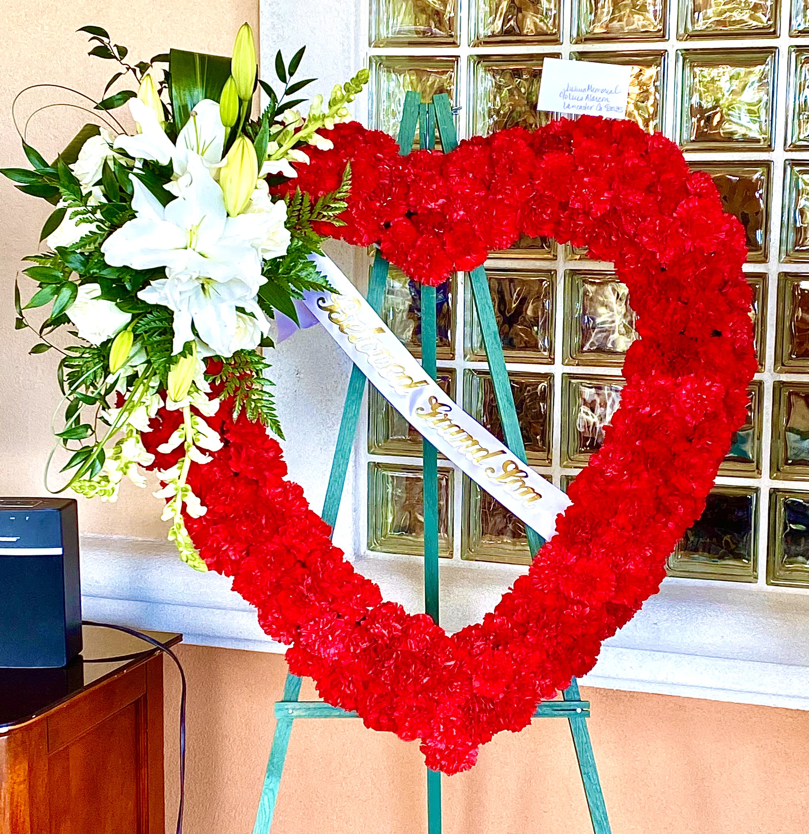 Heart-shaped red floral wreath on an easel with white lilies and a ribbon