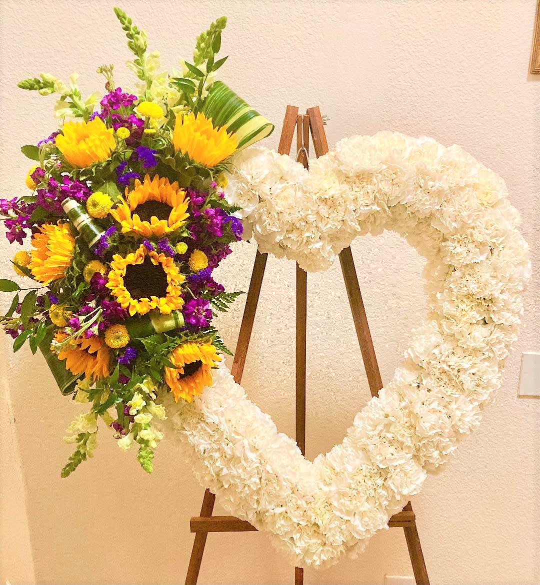 Heart-shaped white floral wreath on an easel beside a colorful sunflower bouquet.