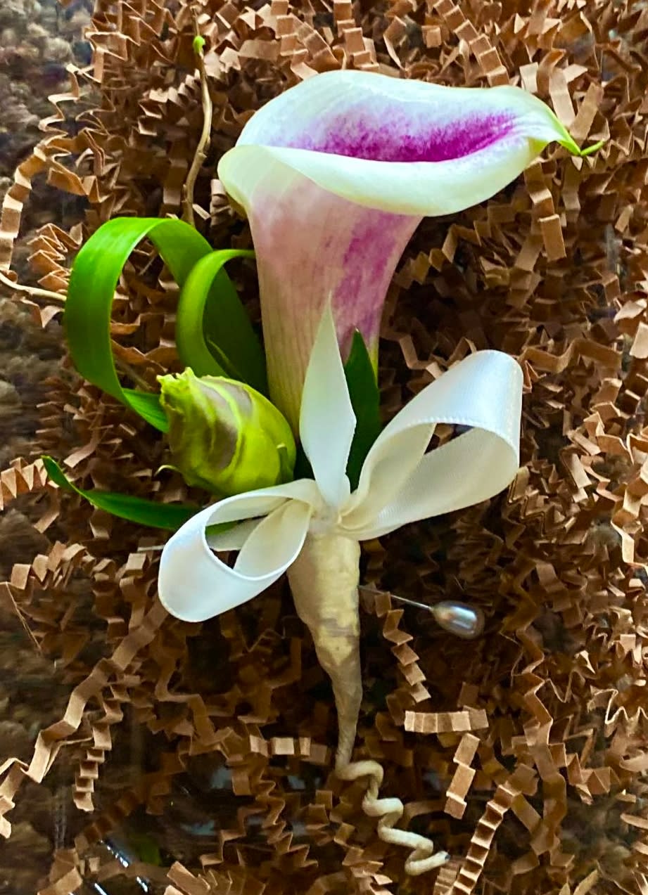 A calla lily boutonniere with white ribbon on brown filler