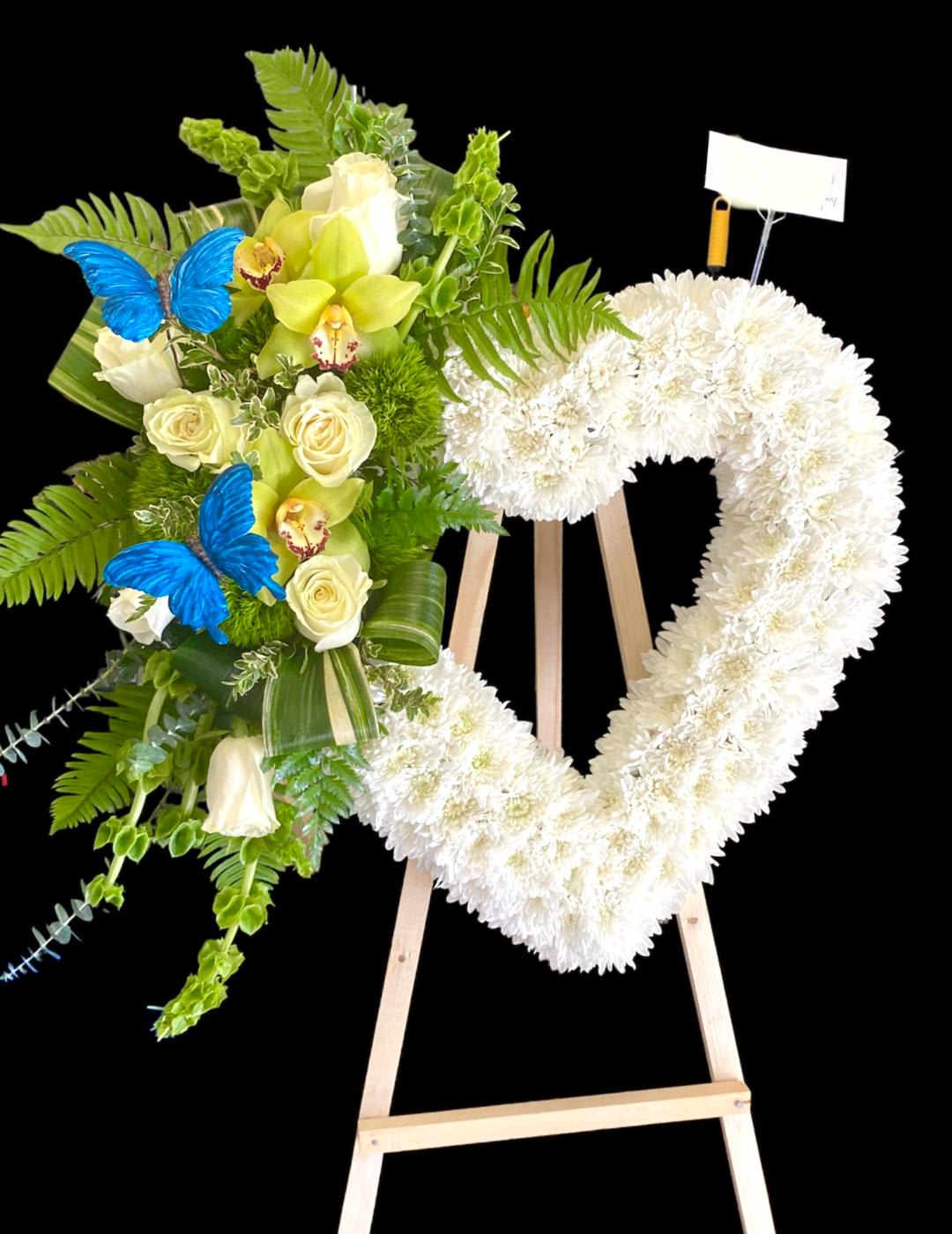 Heart-shaped white floral wreath on an easel beside a green and white bouquet with blue butterflies