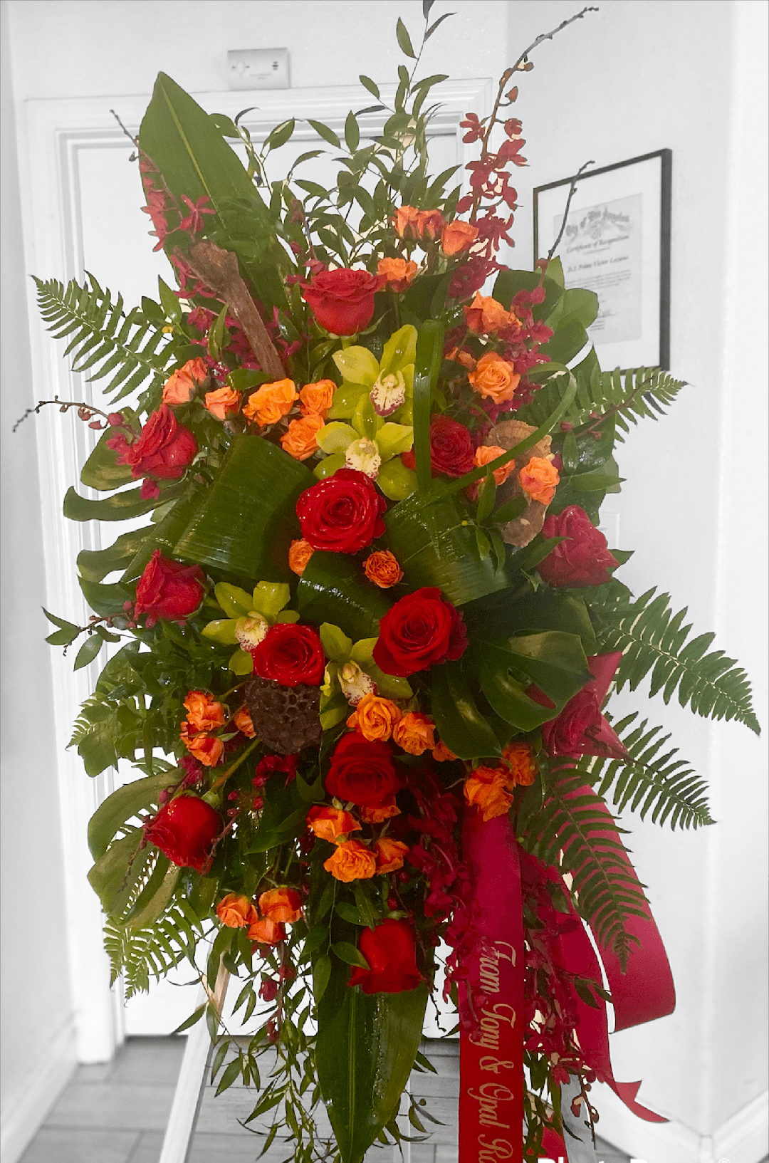 Rose &amp; Orchid Sympathy Standing Spray  - This vibrant sympathy tribute includes a variety of blooms including Ecuadorian Red Roses, Orange Spray Roses, Green Cymbidium Orchids, &amp; Ruby Red Mokara Orchids. All set in various lush foliages. This design measures approx. 28" W x 48" H. Includes personalized ribbon. 