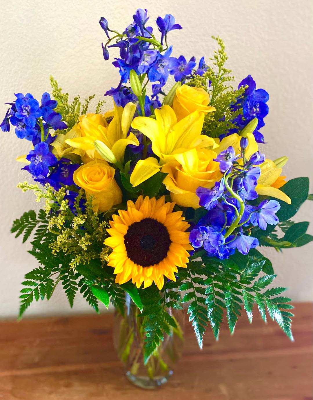 Yellow roses, lilies, and a sunflower in a glass vase with blue flowers