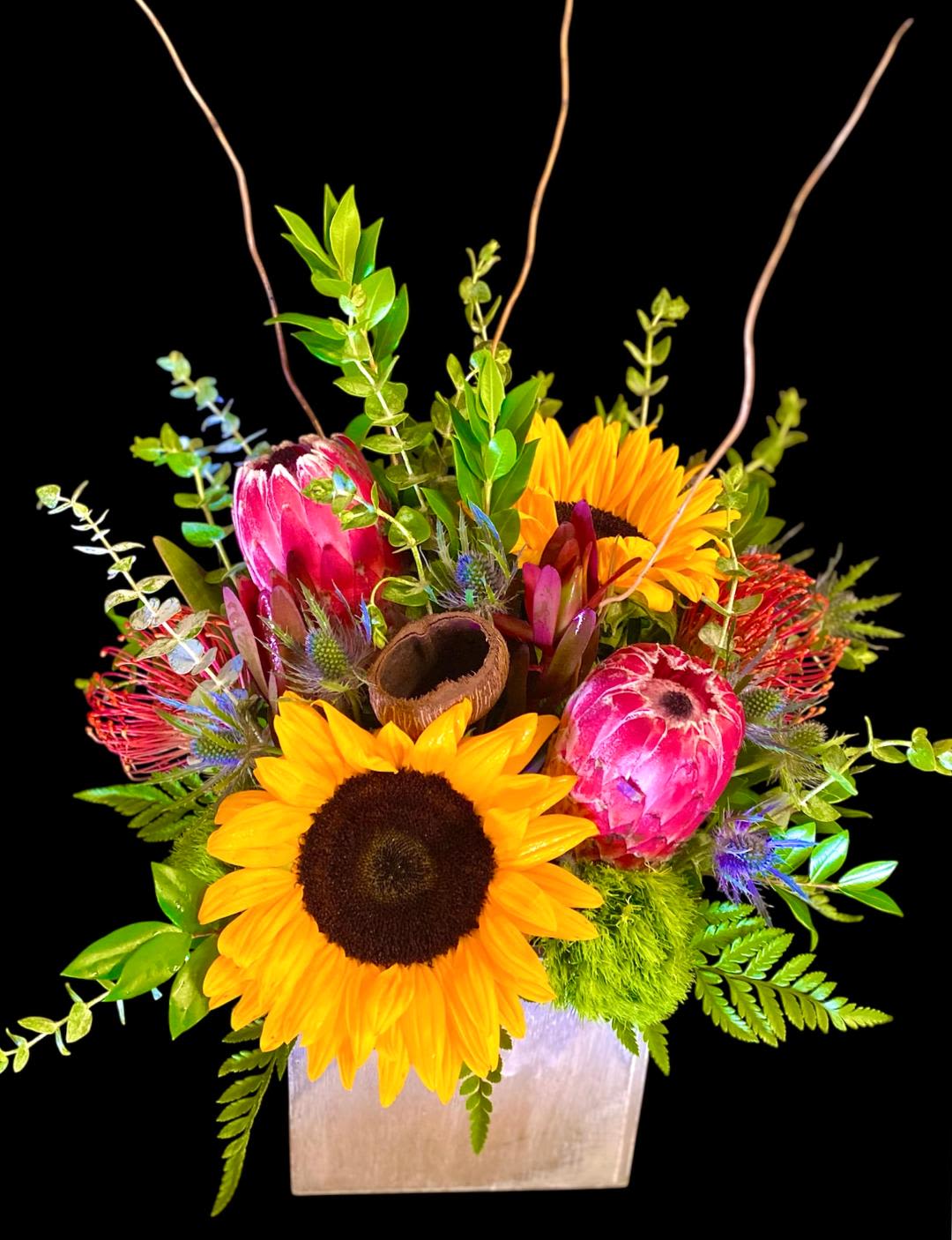 Sunflowers and pink protea in a square vase with greenery
