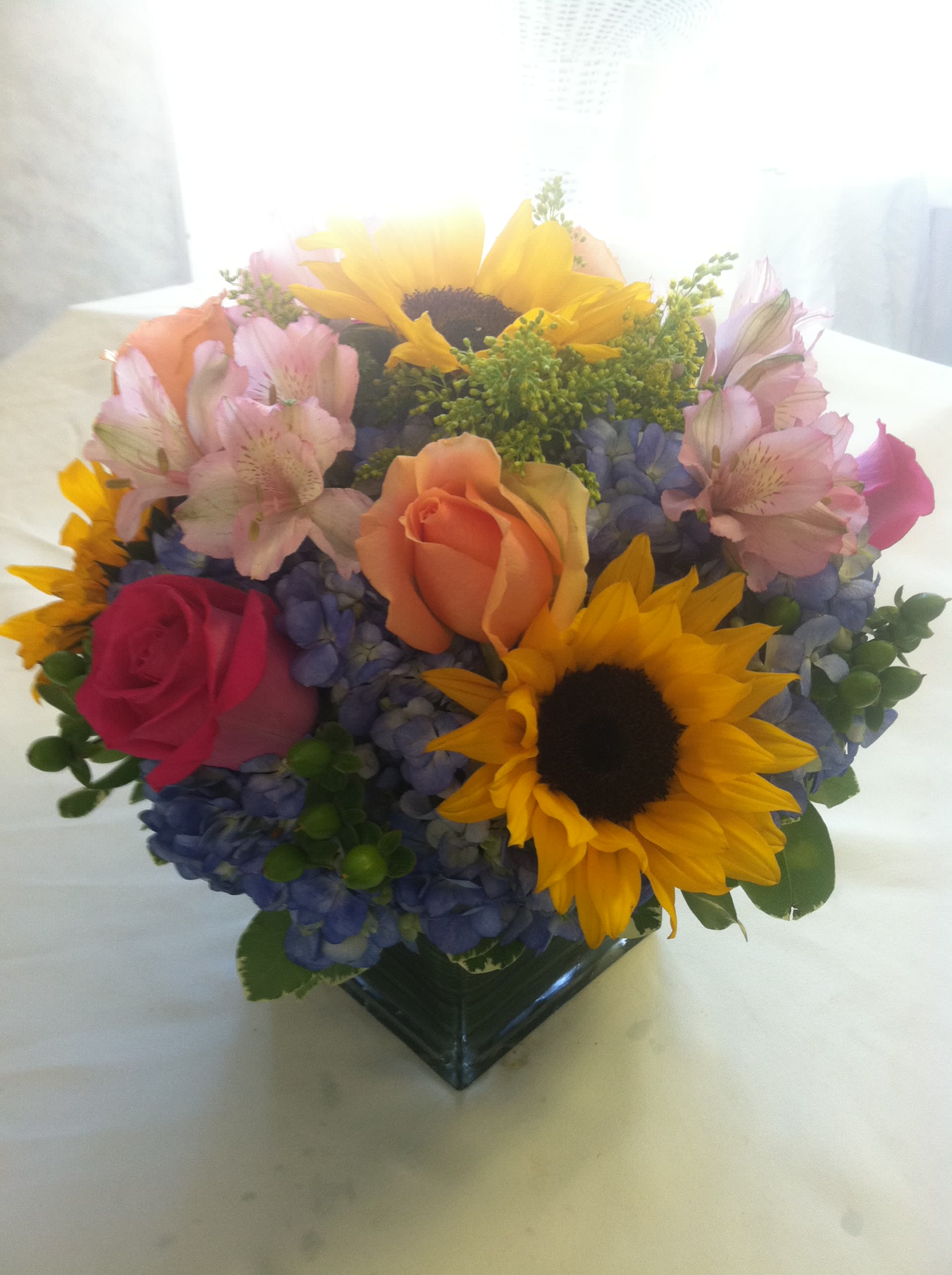 Lush and Beautiful Centerpiece - This fun summer themed centerpiece is great for parties, weddings, and even can be used as a gift for any special occasion! Roses, hydrangea, sunflowers, and more make a wonderful bright bouquet. Hand-crafted and designed in a glass cube vase. Perfect for the Summer months!