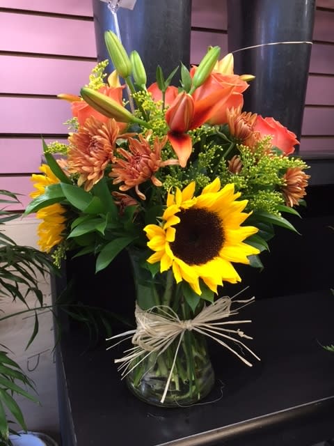 Elegant Autumn Themed Bouquet - Simple, elegant, and warm. Three words that create a great gift for the one you love. This beautiful Autumn bouquet possesses those three attributes and then some. Lilies, Sunflowers, Spider Mums, and more make up this wonderful bouquet. Hand-crafted and designed in a clear glass vase adorned with a raffia bow. Great for any special occasion during the Autumn season. Please Note: Our flowers come in fresh every day. Lilies especially will usually come in bud form. One of the better parts of a bouquet with lilies is watching them bloom right in front of your eyes. A perk to any lily arrangement.