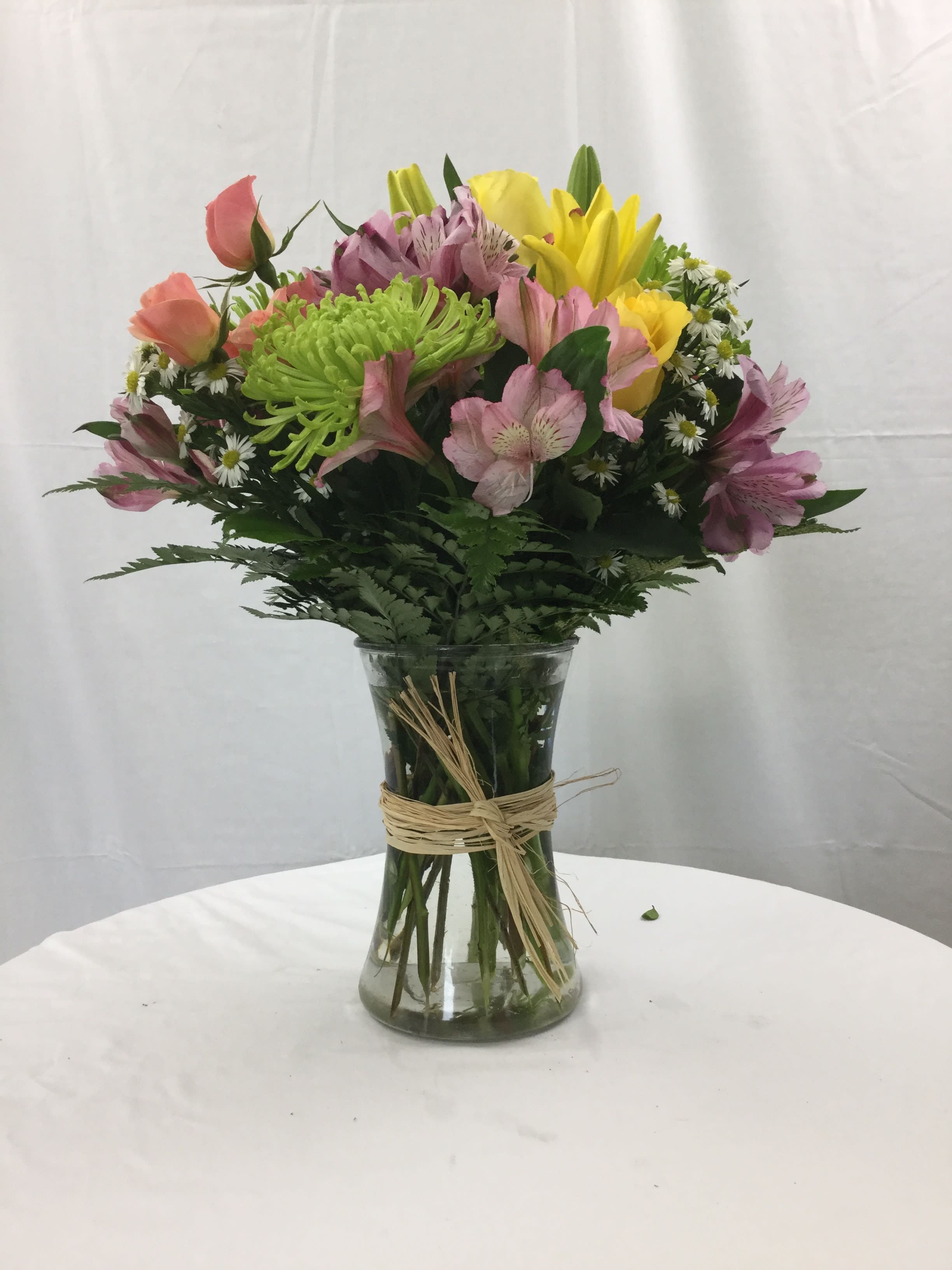 Mixed bouquet of yellow lilies, peach roses, and pink blooms in a clear vase with a raffia tie