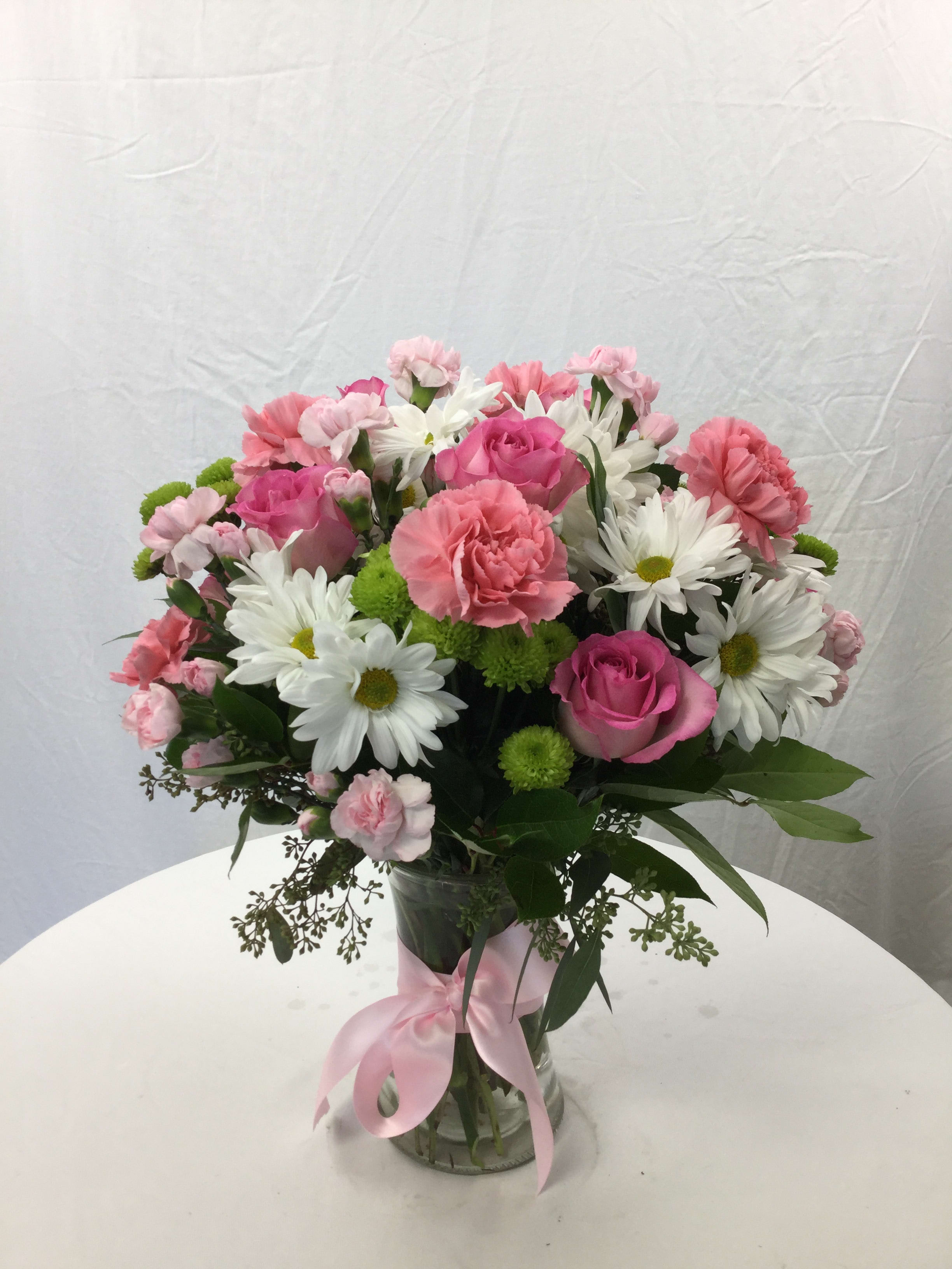 Pink roses, carnations, and white daisies in a glass vase with a pink ribbon.