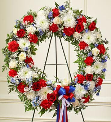 Serene Blessings Red, White &amp; Blue Standing Wreath - This beautiful red, white and blue wreath is a dignified expression of your feelings of love and care for a beloved veteran or patriot. Exquisitely crafted by our expert florists, it features the freshest red and white roses, blue delphinium and more, creating an unforgettable tribute to honor their life. Note: Image shown is Premium.