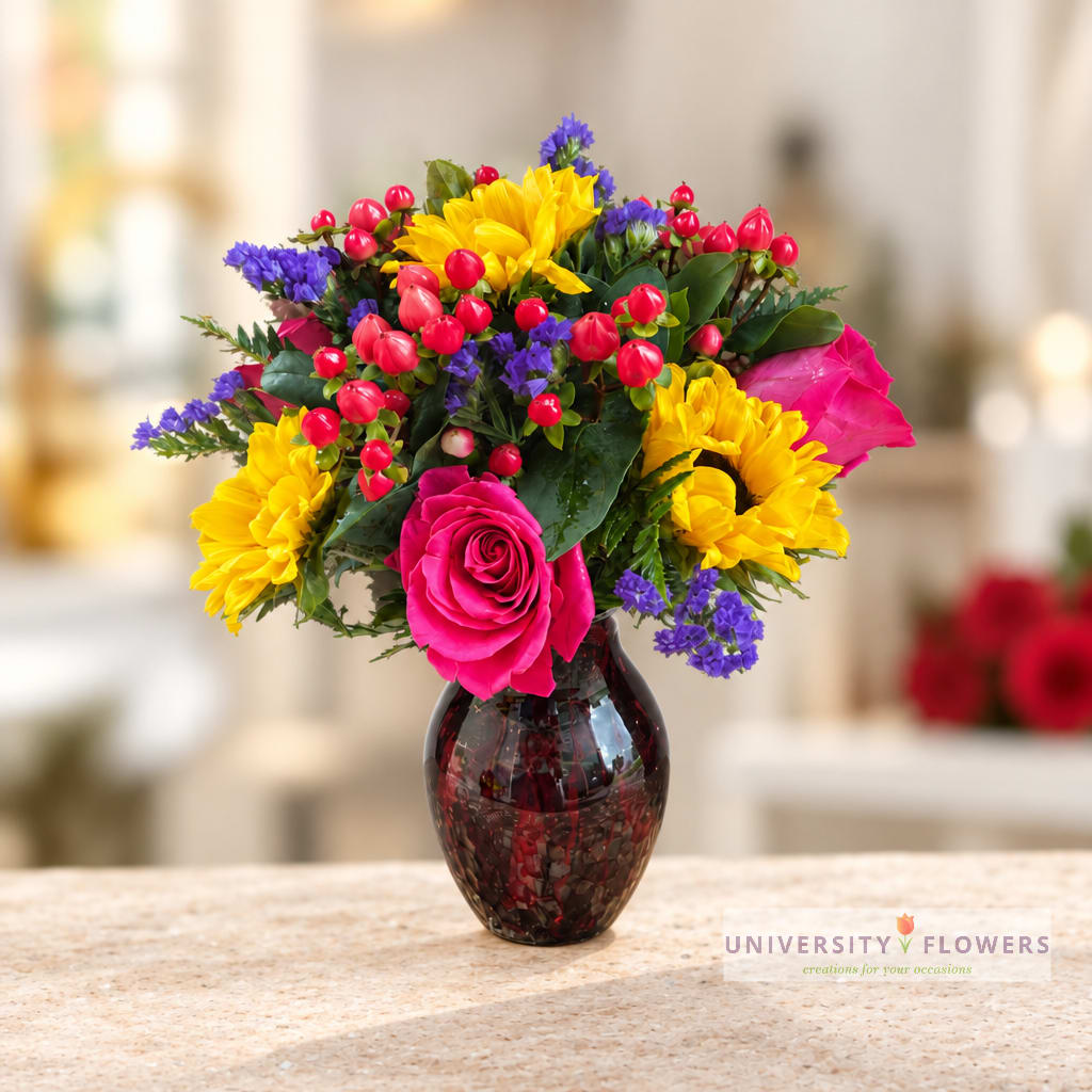 Bright mixed bouquet with pink roses, yellow sunflowers, and purple accents in a dark glass vase