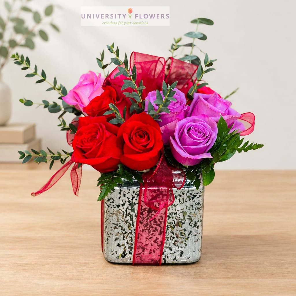 Red and purple roses arranged in a square vase with a pink ribbon